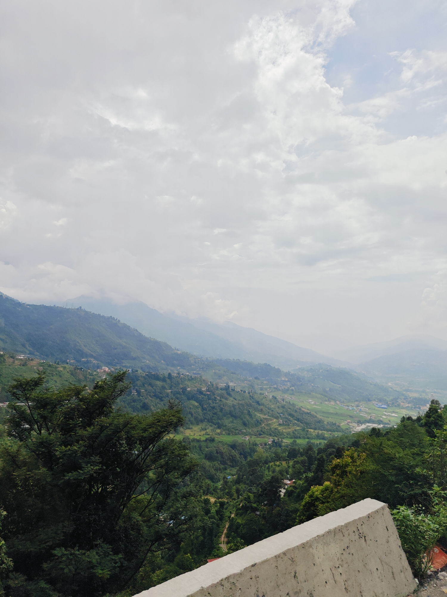 The image depicts a panoramic view of the mountainous terrain surrounding Chandragiri, Nepal. The foreground features a portion of a concrete barrier, likely part of a roadside or viewing platform. Beyond this barrier, a lush, green landscape unfolds with rolling hills covered in dense vegetation and scattered settlements.
The middle ground is dominated by terraced fields and the winding roads that navigate the steep slopes. Small villages, identifiable by their clusters of buildings, are visible nestled within the valleys and on the hillsides. The architecture appears to be traditional, with muted colors that blend into the natural environment.
In the background, a series of mountains recede into the distance, their peaks partially obscured by a soft, atmospheric haze. The sky above is overcast with a thick layer of clouds, suggesting a humid or possibly rainy day. The overall impression is one of natural beauty and rural tranquility, characteristic of the Himalayan foothills. The scene is devoid of any people, animals, or specific activities, focusing solely on the landscape and its man-made elements.