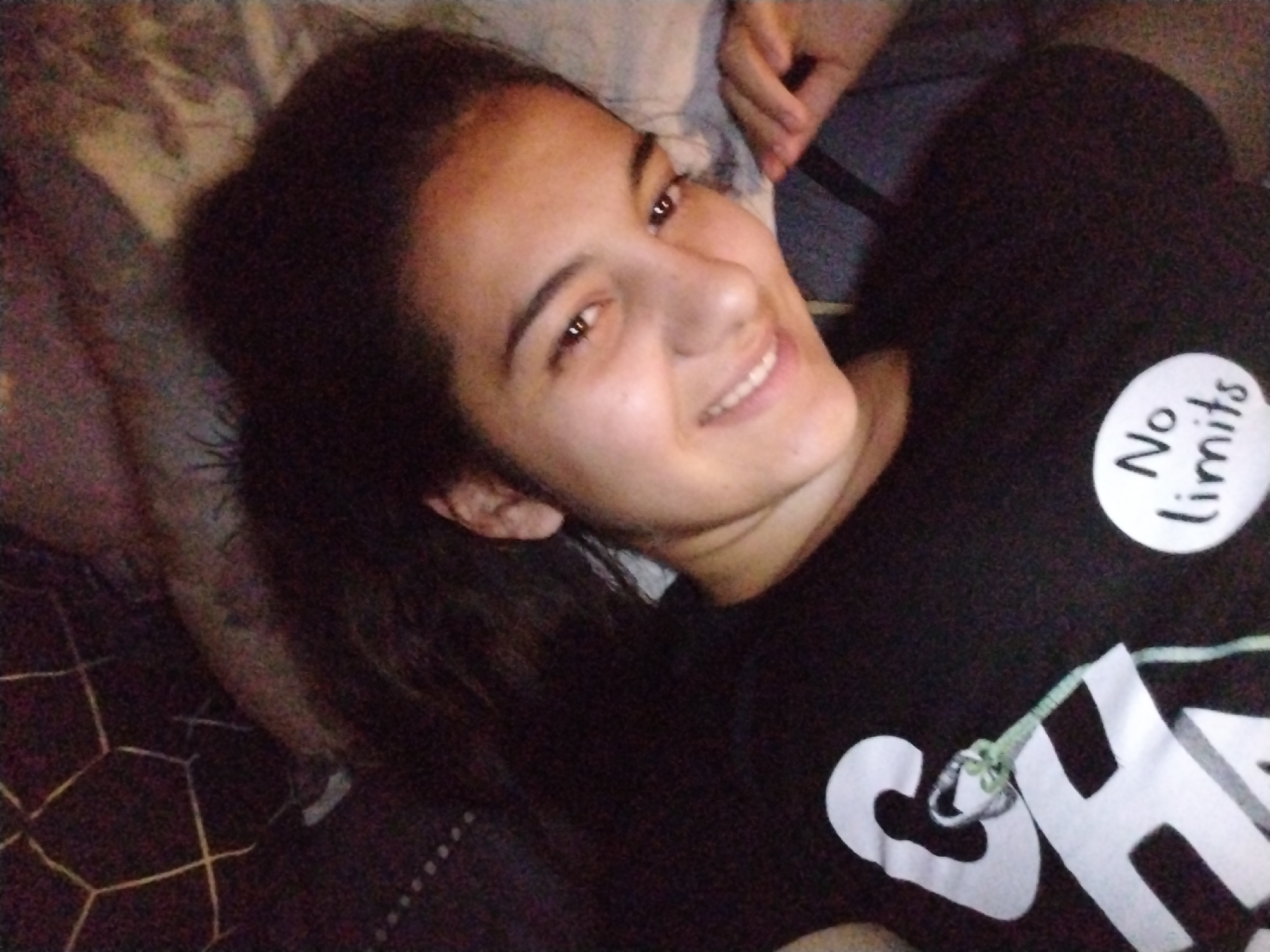 The image shows a young woman lying down and smiling directly at the camera. She has long, dark hair, possibly styled in a ponytail, which rests on a surface behind her head. Her expression is happy and relaxed, with a wide smile showing her teeth. She is wearing a black t-shirt. A white, circular badge is pinned to her upper chest, displaying the text "No limits" in black cursive script. Below this badge, the white letters "CH" are partially visible on the t-shirt, alongside what appears to be part of another letter, and a light green or teal-colored strap with a small metallic ring and plastic elements is draped over this area. In the upper right corner, a hand is visible, resting on a dark surface. The background is mostly blurred, showing soft, textured fabrics, one section of which features a dark geometric pattern with lighter lines. The lighting is somewhat dim, suggesting an indoor setting, possibly an evening or low-light environment. The overall impression is one of casual comfort and contentment.