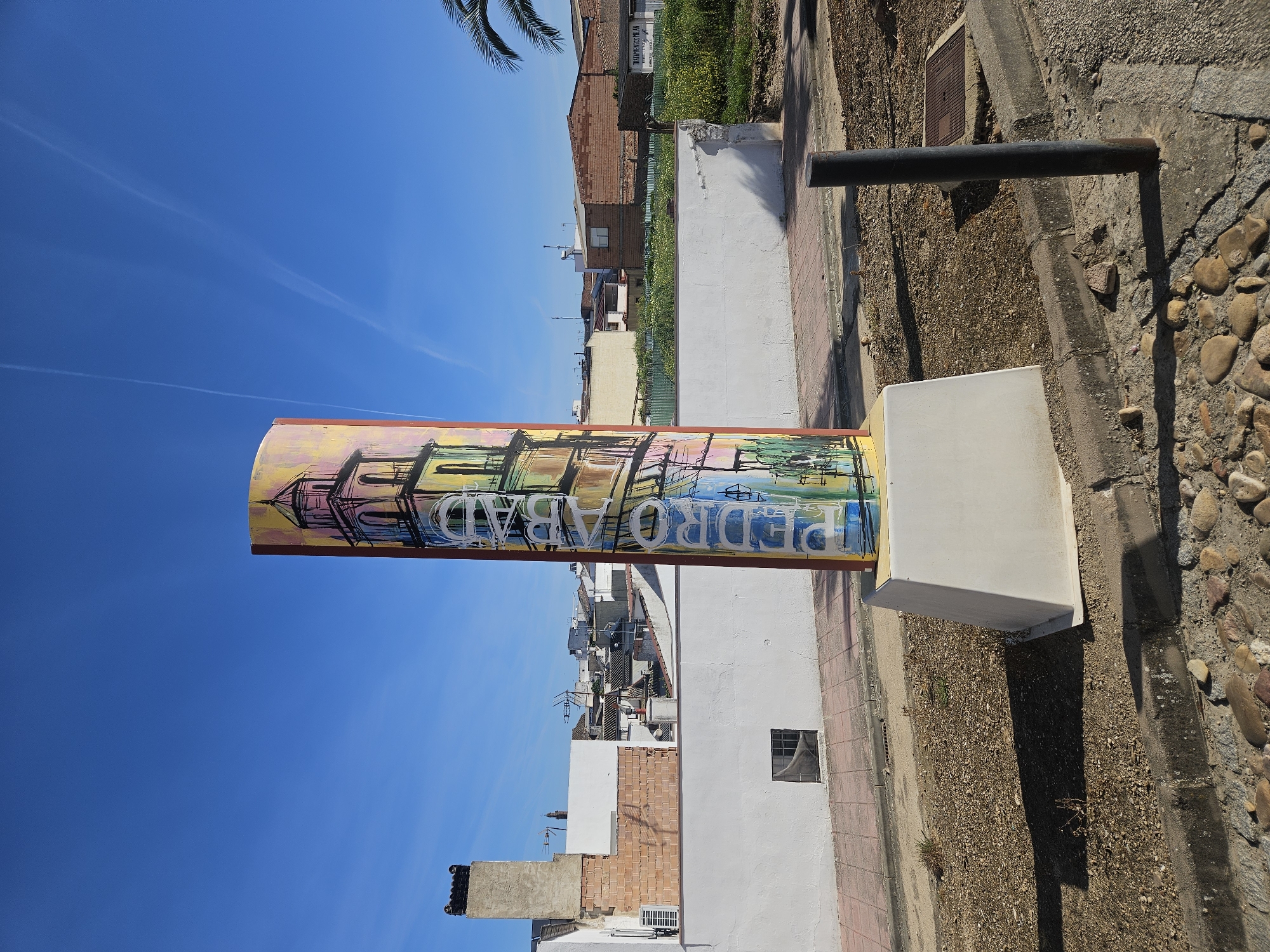 The image shows a vertical sign in Pedro Abad, Spain, that reads "PEDRO ABAD" in stylized white letters against a backdrop of colorful illustrations resembling buildings. The sign is mounted on a white square pedestal. A black metal bar is positioned near the sign. The environment includes buildings, a sidewalk, and a patch of ground covered with gravel and rocks. The sky is clear and blue.