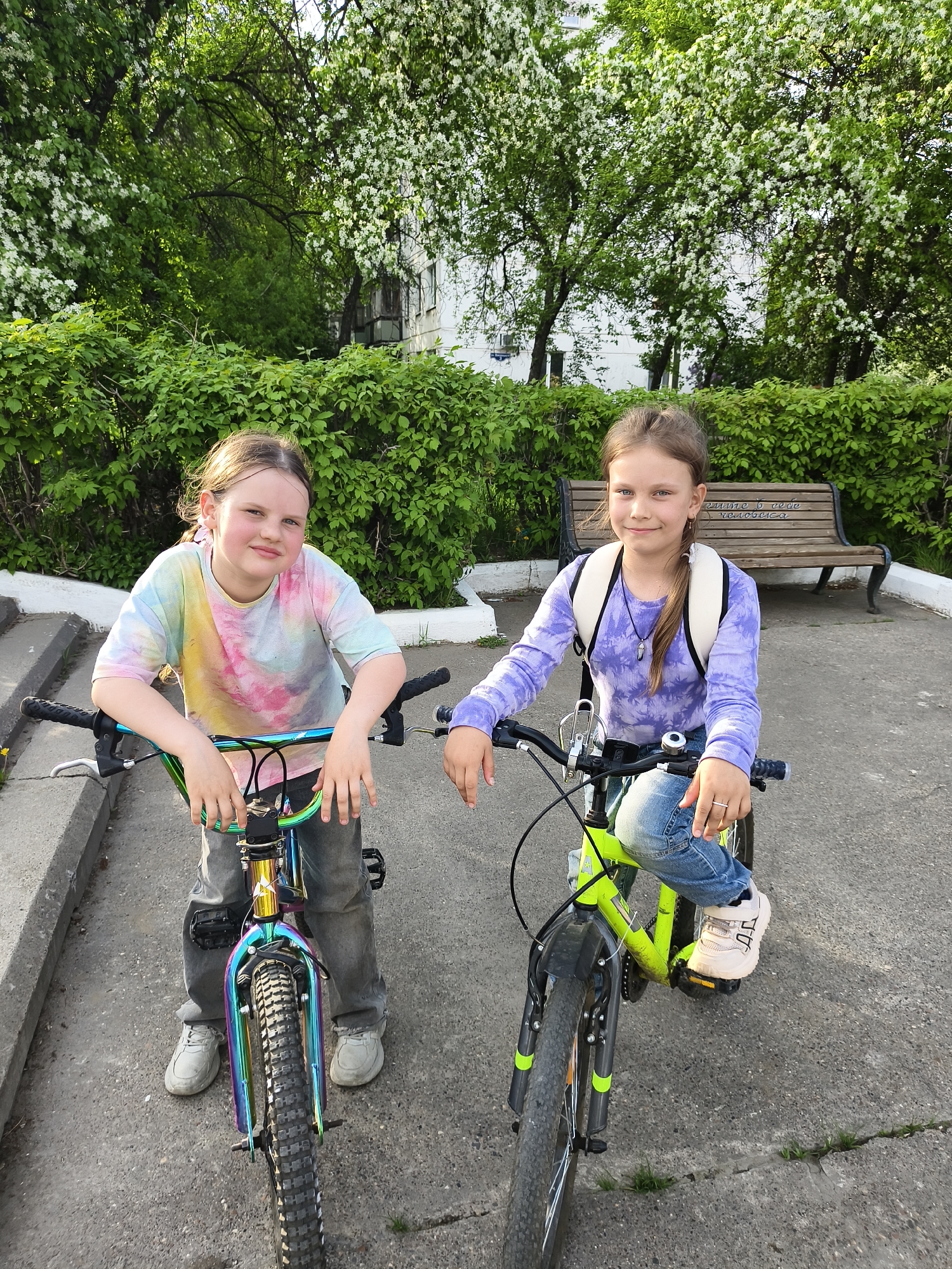 Two young girls stand with their bicycles on a paved surface in what appears to be a city park setting. The girl on the left has her hands resting on the turquoise handlebars of a bicycle that has a multicolored frame and black tires. She is wearing a tie-dye t-shirt and grey jeans with white sneakers. The girl on the right is seated on a lime green bicycle, her right foot on a pedal. She wears a purple long-sleeved top, blue jeans, and white sneakers, with a white backpack slung over her shoulders. Behind the girls is a bench with writing on it, situated in front of a dense green hedge. Flowering trees are visible in the background, suggesting a park or garden environment. The lighting suggests it's daytime.