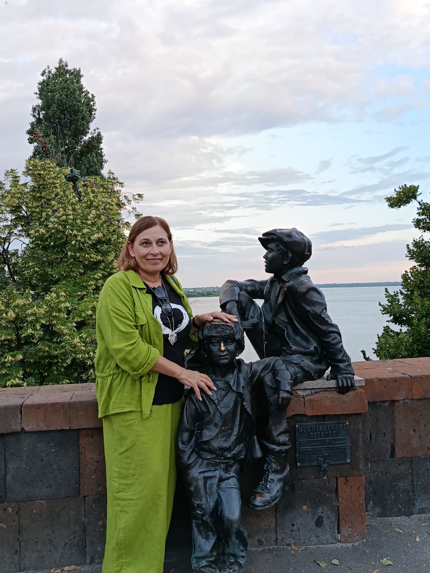 A woman, with medium-length brown hair and wearing a bright green jacket and matching pants, stands to the left of two bronze statues of young boys. Her left hand rests on the shoulder of the statue in front, while her right hand is on the knee of the statue behind. The statues are positioned on a low, brick wall overlooking a body of water. The statue in the foreground is seated, while the statue behind it is leaning against the wall. Behind the statues and the woman are trees and a cloudy sky. A plaque is attached to the wall below the statues. This is the Monument to Childhood, located on the embankment of Saratov, Russia.