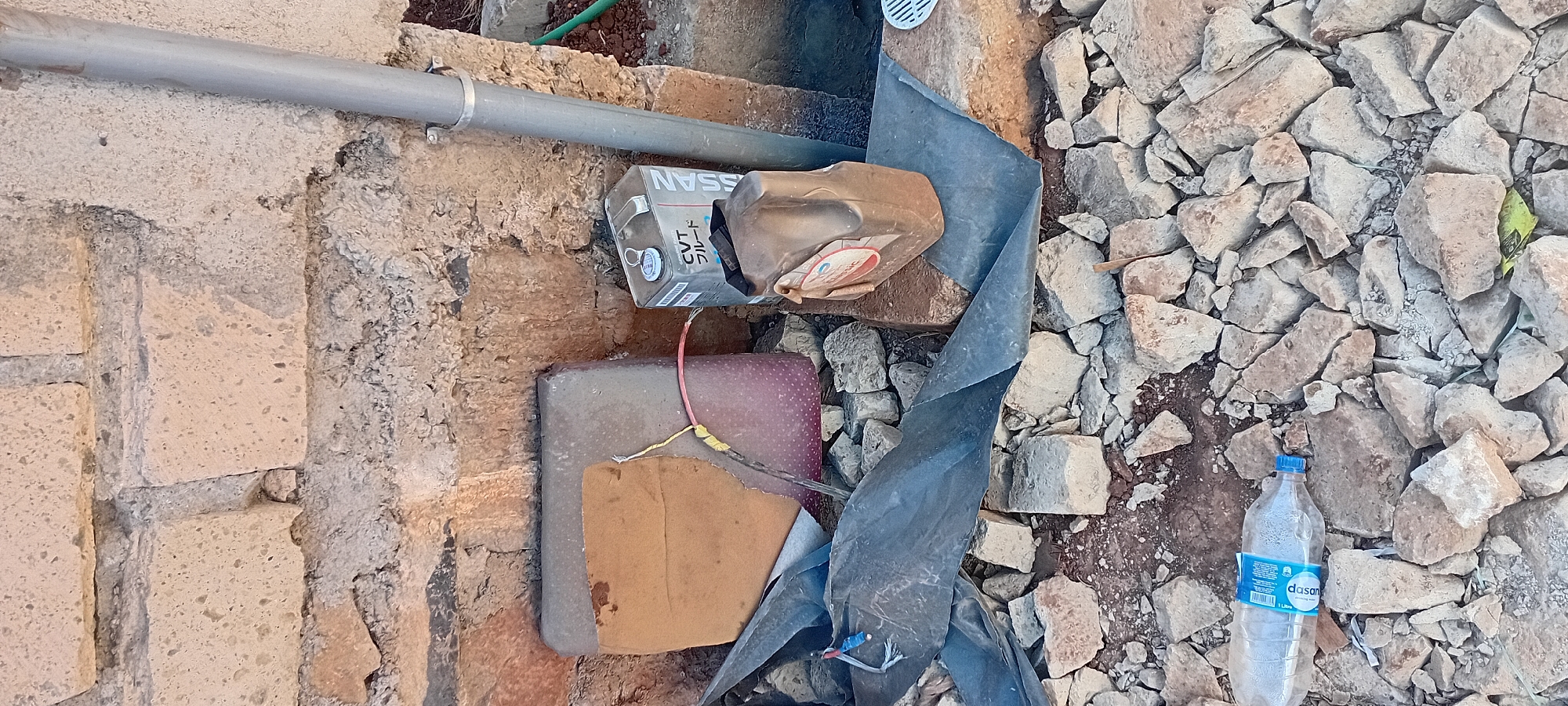 The image depicts an outdoor scene with a focus on discarded items and construction debris. In the foreground, a clear plastic water bottle lies on a bed of broken stones and rubble. The bottle appears to be partially filled with water and has a blue cap. Behind the bottle, a black plastic sheet is crumpled and partially covers more stones.
In the middle ground, there are two plastic containers and a metallic can. The metallic can, positioned upright, is silver with a black and white label that reads "CVT" and Japanese characters. It has a silver cap. To the left of the can is a large, rectangular object, possibly a container or a block, with a reddish-pink speckled surface and a brown material covering its top. A red string is tied around this object, connecting to something off-frame. To the right of the metallic can, a brown, opaque plastic container is angled on its side, with a partially visible circular label featuring red and white text.
The background consists of a rough, textured brick wall made of light-colored, porous bricks. A section of a grey pipe runs vertically along the right side of the wall, secured by a metal clamp. The ground beneath the wall and the objects appears to be a mixture of dirt and more broken stones, indicating a construction or demolition site. The overall environment suggests an area in Kijabe Ward, Kenya, characterized by unfinished or damaged structures and accumulated waste. There are no people visible in the image.