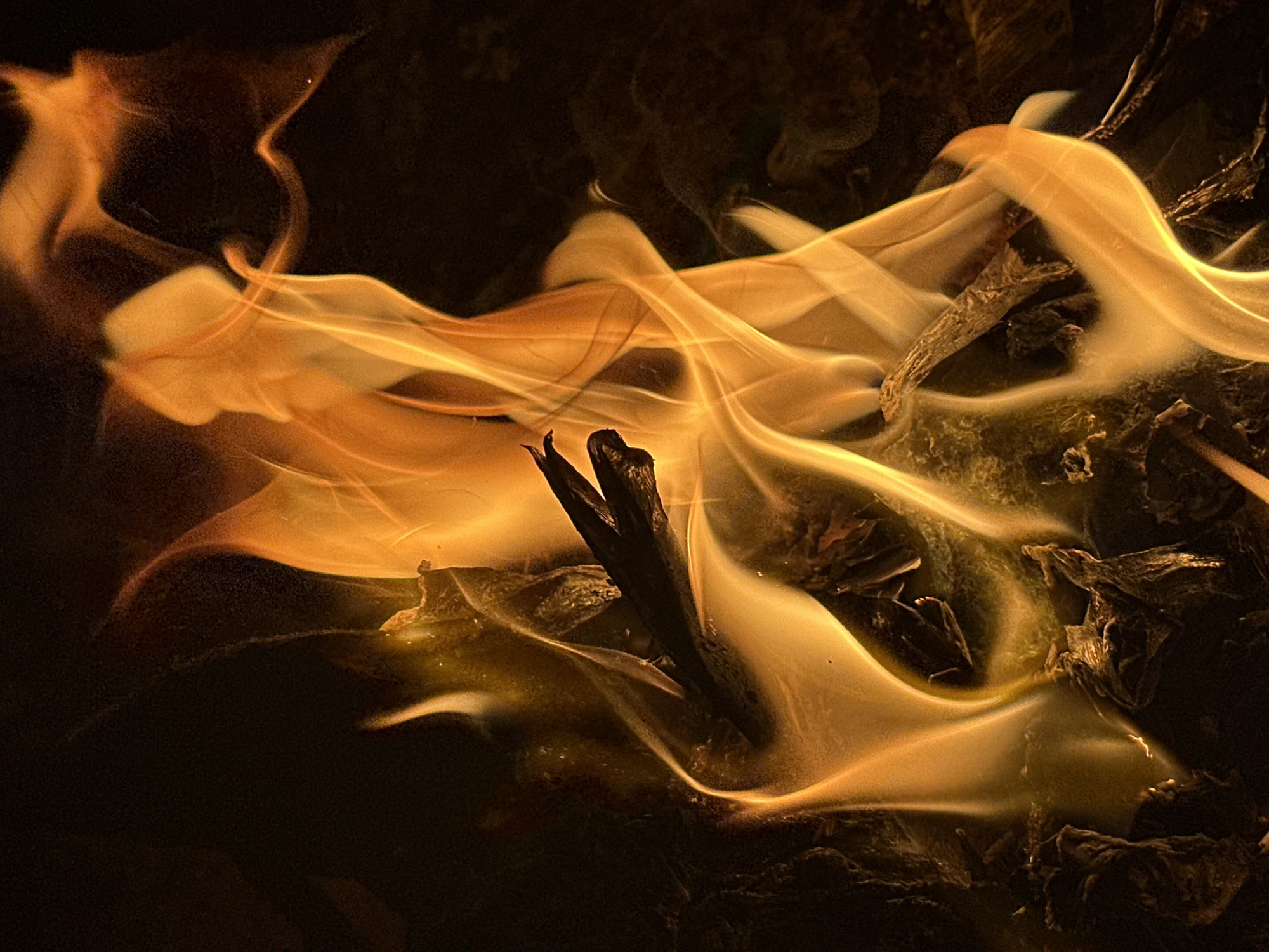The image shows a close-up of a fire burning in Bar Gada, Nigeria. The flames are predominantly orange and yellow, with visible smoke. Charred wood and debris are visible within and around the flames. A small, dark stick is prominent in the center of the fire. The surrounding environment is dark. There are no people visible.