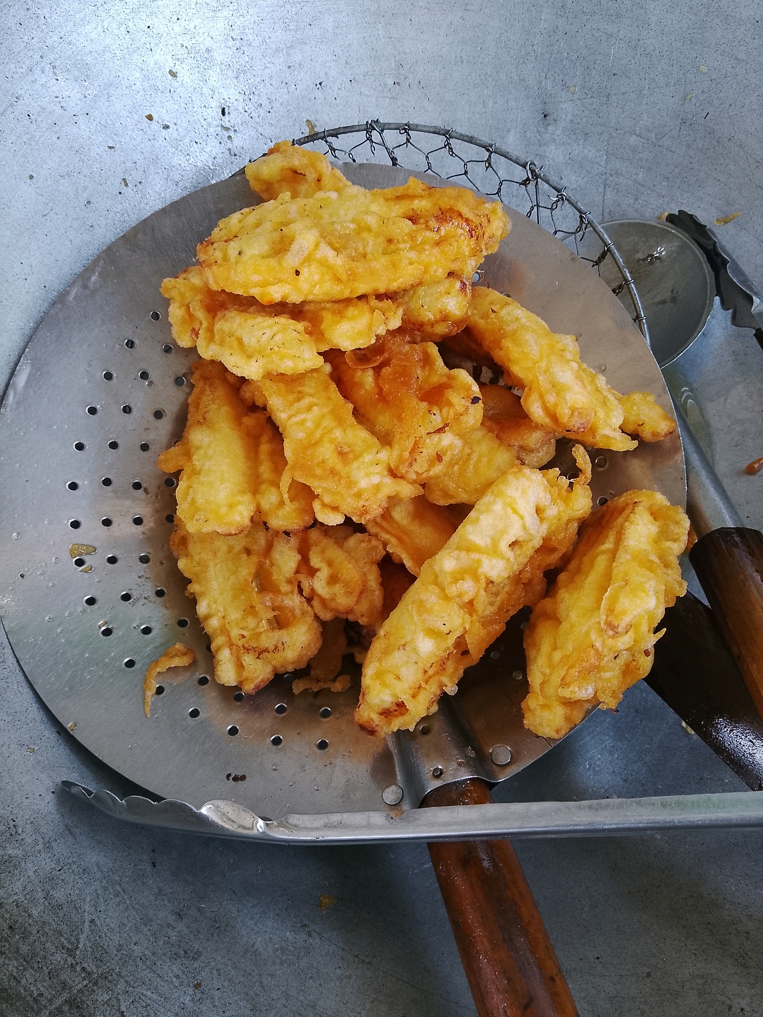 In MalangSari, Indonesia, a close-up, overhead shot captures a metal strainer filled with golden-brown, deep-fried fritters resting on a textured, grey surface. The fritters, appearing to be bananas or a similar fruit coated in batter, are irregularly shaped and piled atop each other. They are slightly oily, indicating they were recently fried. The strainer itself is round with numerous small holes and is supported by two wooden-handled utensils, one on the left and one on the right, which are also resting on the grey surface. The background is a continuation of the grey, textured surface, showing some minor imperfections and specks. A small, metallic object, possibly a lid or another cooking utensil, is partially visible to the right of the strainer. The lighting is even, with no harsh shadows, suggesting an indoor setting.