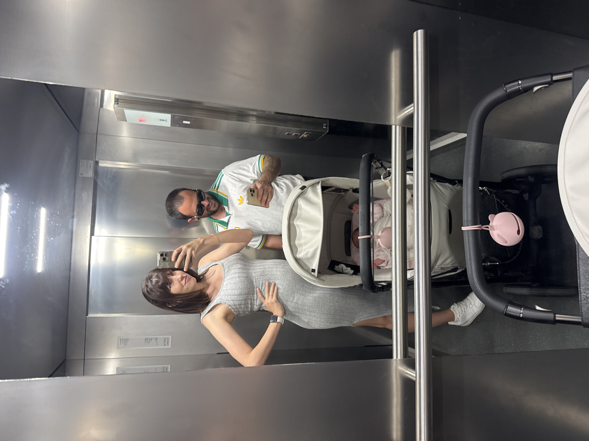 A couple in an elevator with a baby stroller are taking a selfie in a mirror. The woman is wearing a gray dress and white sneakers. The man is wearing a white soccer jersey with green and yellow accents and sunglasses. The stroller is white with a black frame. The elevator appears to be made of stainless steel. It is not possible to ascertain the location in Pomezia, Italy from the image.