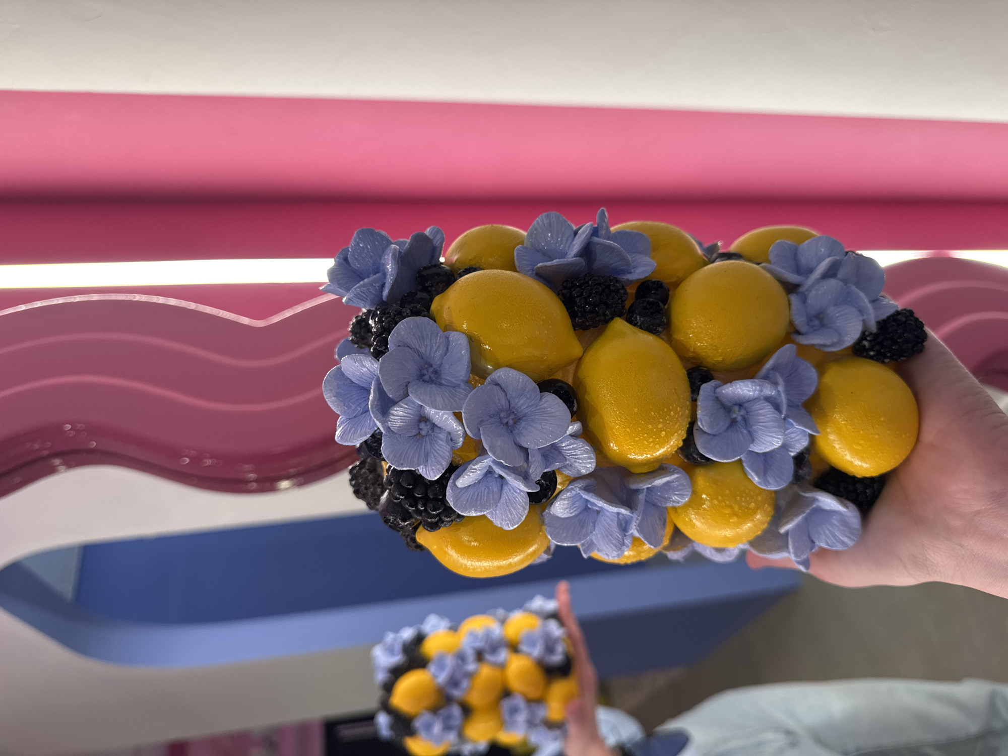 The image shows a person holding a decorative arrangement consisting of yellow lemons, dark blackberries, and periwinkle hydrangea flowers. The arrangement is compact and possibly part of a larger display or decoration.
In the background, there is a colorful interior design element featuring a combination of pink, white, and blue wavy patterns. The scene is brightly lit, suggesting indoor lighting. Another similar decoration appears blurred in the lower portion of the image.
Although there are details provided in the request, there is no information in the image to verify that this scene took place in the city of Sochi, Russia.