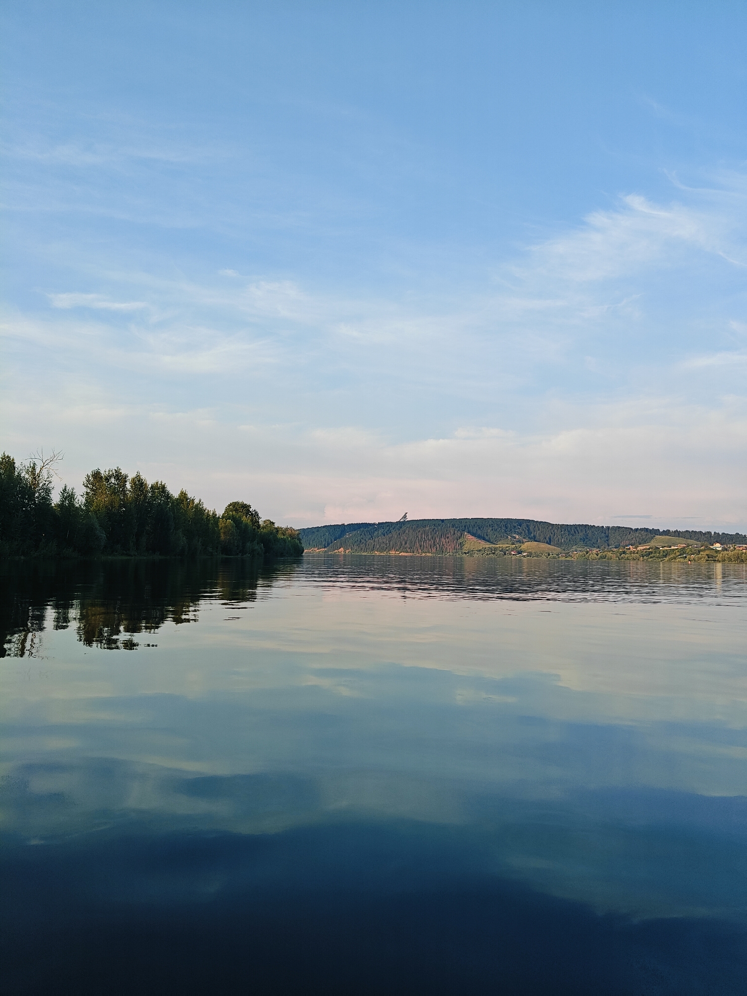 The image depicts a serene natural landscape with a wide river in the foreground and a forested, hilly background. The sky is a pale blue with wispy white clouds. The river's surface is calm, reflecting the sky and the trees on the opposite bank. The left bank of the river is lined with a dense growth of deciduous trees. The right bank features rolling hills covered in pine trees and scattered buildings. The overall impression is one of tranquility and natural beauty, typical of a rural setting. It is highly probable that this image was captured near the village of Ol'khovskoye in Russia, given the characteristic scenery of the region.