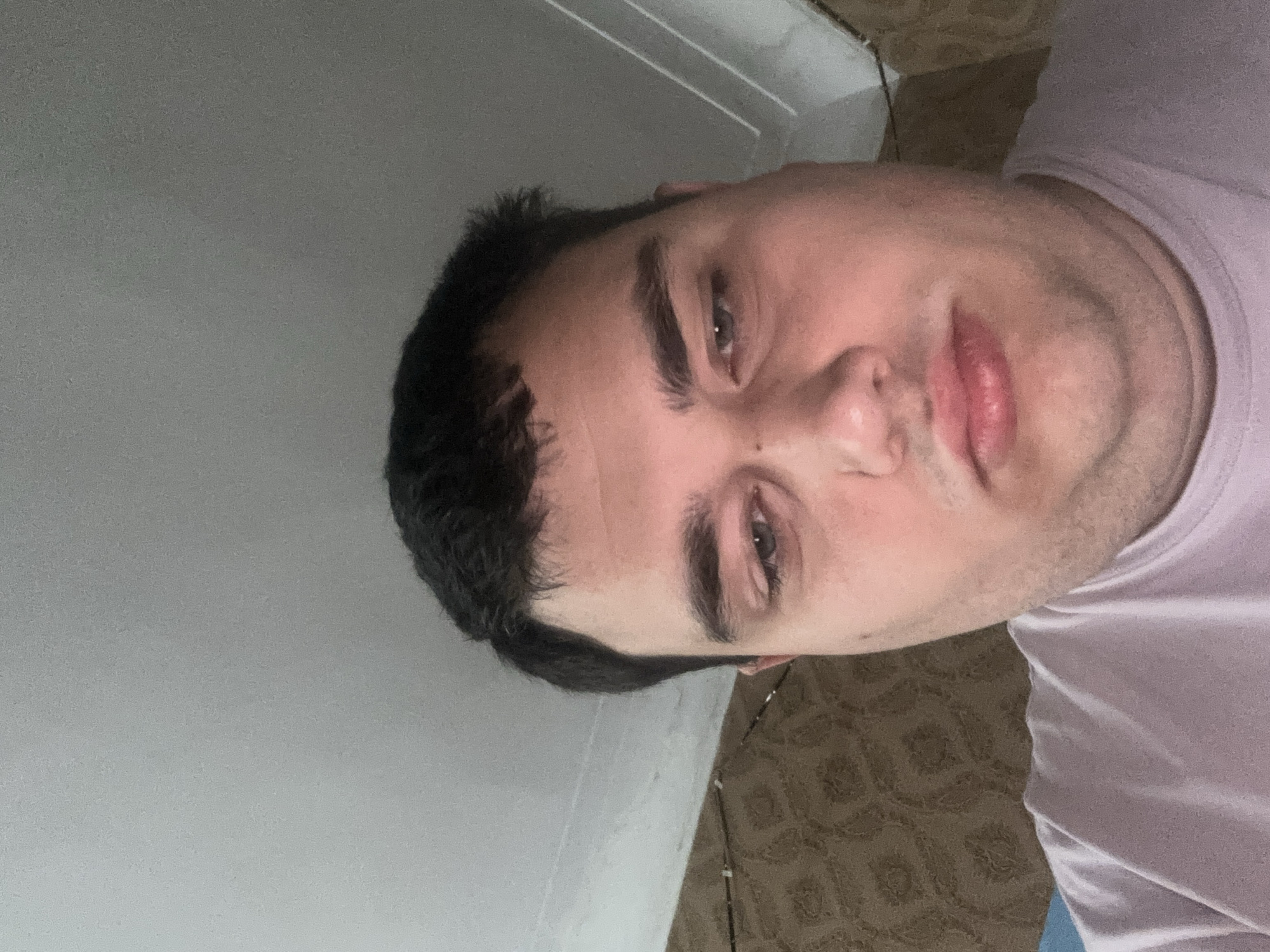 A close-up, overhead shot shows a light-skinned person with dark hair and blue eyes. The person appears to be looking down, with a tired or sad expression. The background consists of a beige patterned wallpaper and a white ceiling with a light fixture. The person is wearing a light purple t-shirt. The setting appears to be indoors, possibly a bedroom.