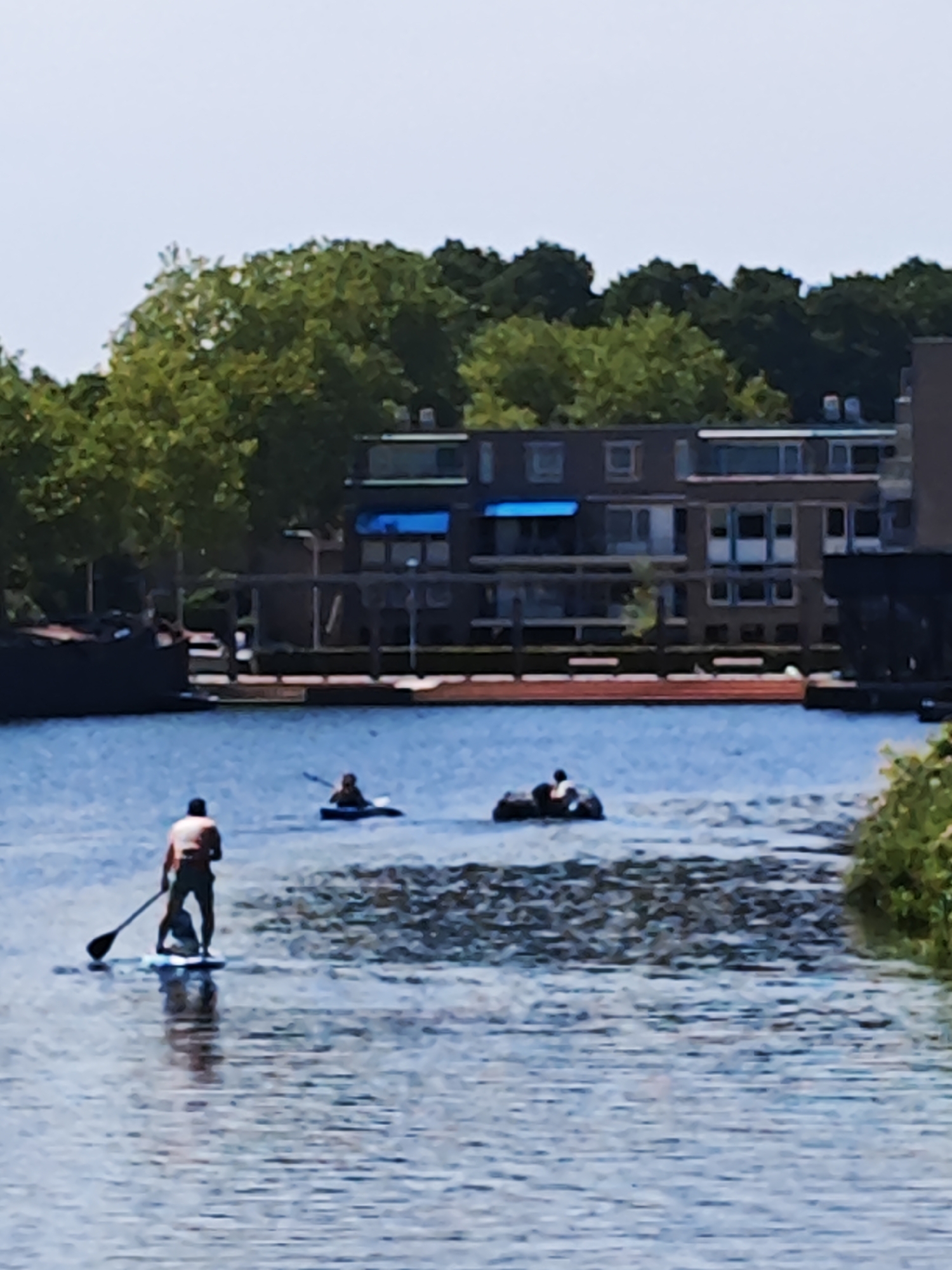 A man is paddleboarding on a body of water in front of a multi-story building. Another person is in a kayak on the water in the background, and a third person is also on the water in a small boat. The water appears calm, and the sky is overcast. Trees line the shore in the background. The scene is likely in Tilburg, Netherlands, given the architectural style of the building and the presence of canals.