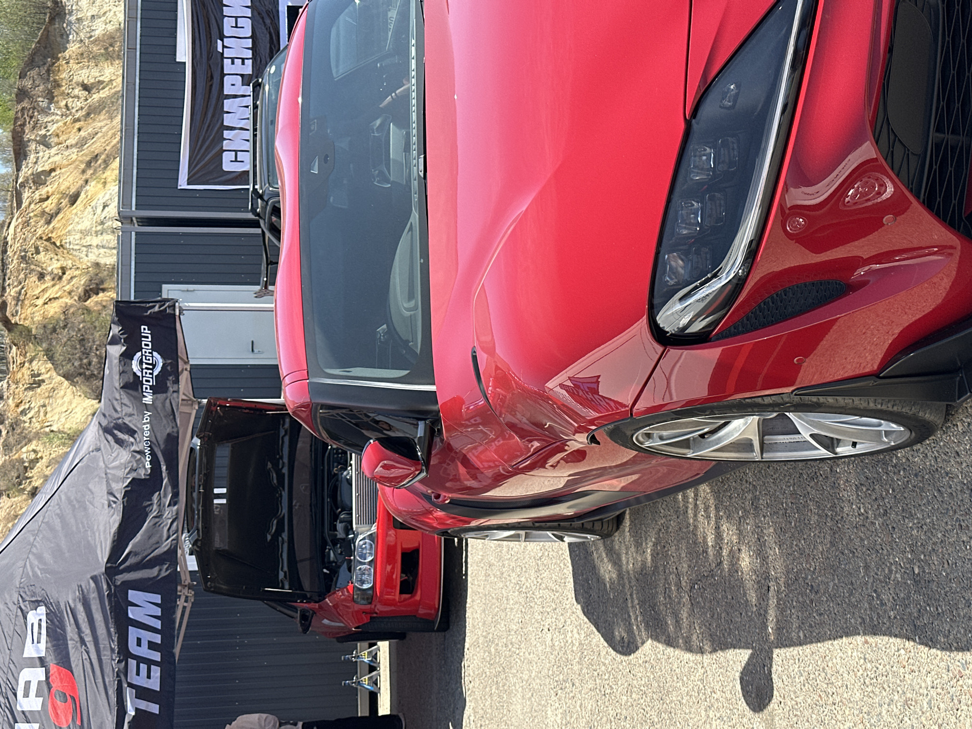 The image shows a close-up view of the front of a red Toyota Supra parked outside a building in Sadovoe, Russia. The car is facing towards the right of the image. Another red car is visible behind it with its hood open. A black tent with white text "TEAM" and "Powered by IMPORTGROUP" is to the left of the cars. A building with a banner containing Cyrillic script is located behind the tent. A cliff face is visible in the background.