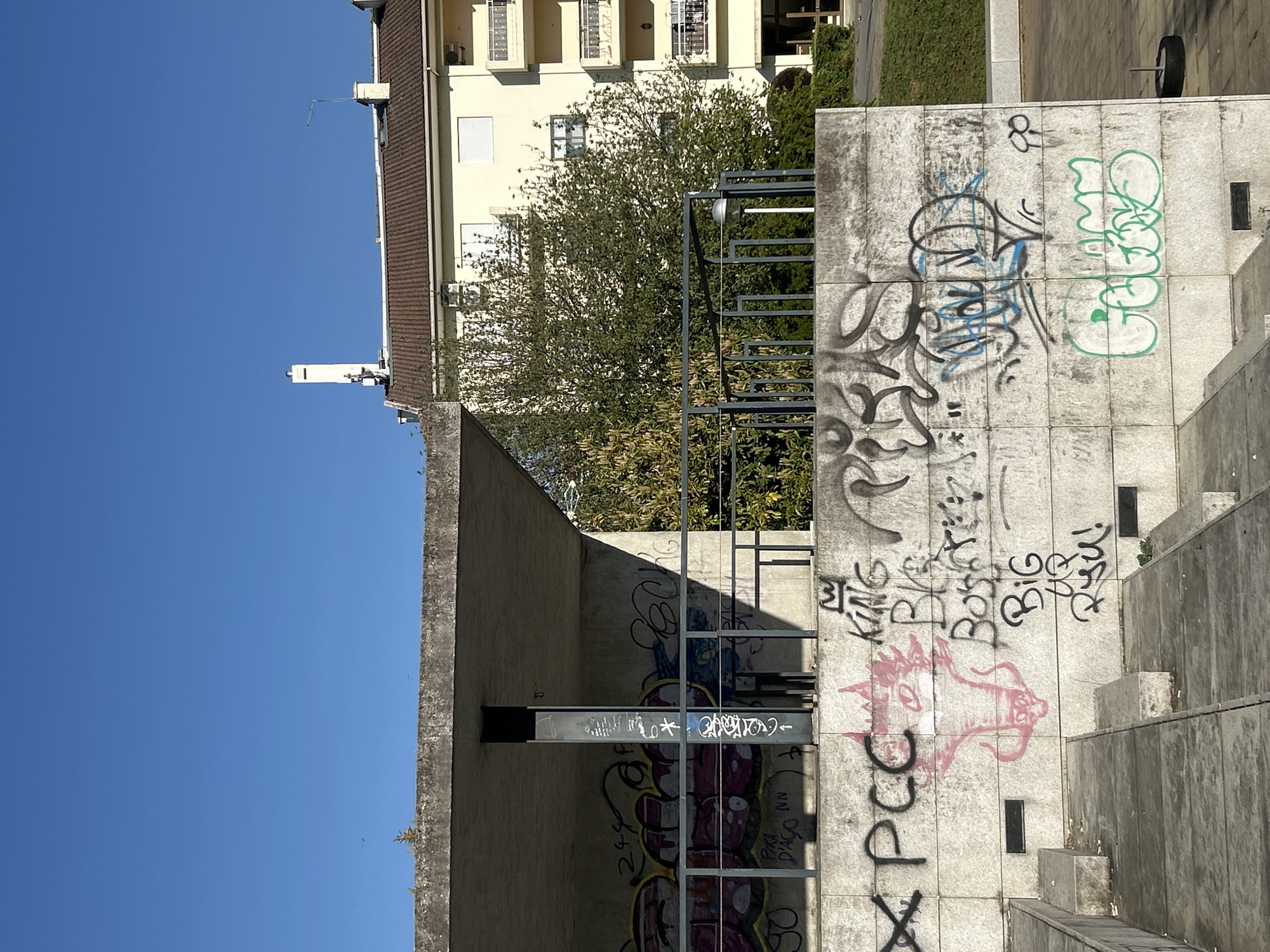 The image depicts an urban scene in Vila Real, Portugal. A concrete structure covered in graffiti dominates the foreground. Adjacent to this structure are concrete steps leading upwards. Behind the graffiti-covered concrete is a gray metal structure. In the background, a multi-story building with a brown roof and beige facade is partially visible, interspersed with greenery from trees. A clear blue sky is overhead. A spare tire is on top of the graffiti-covered structure.
