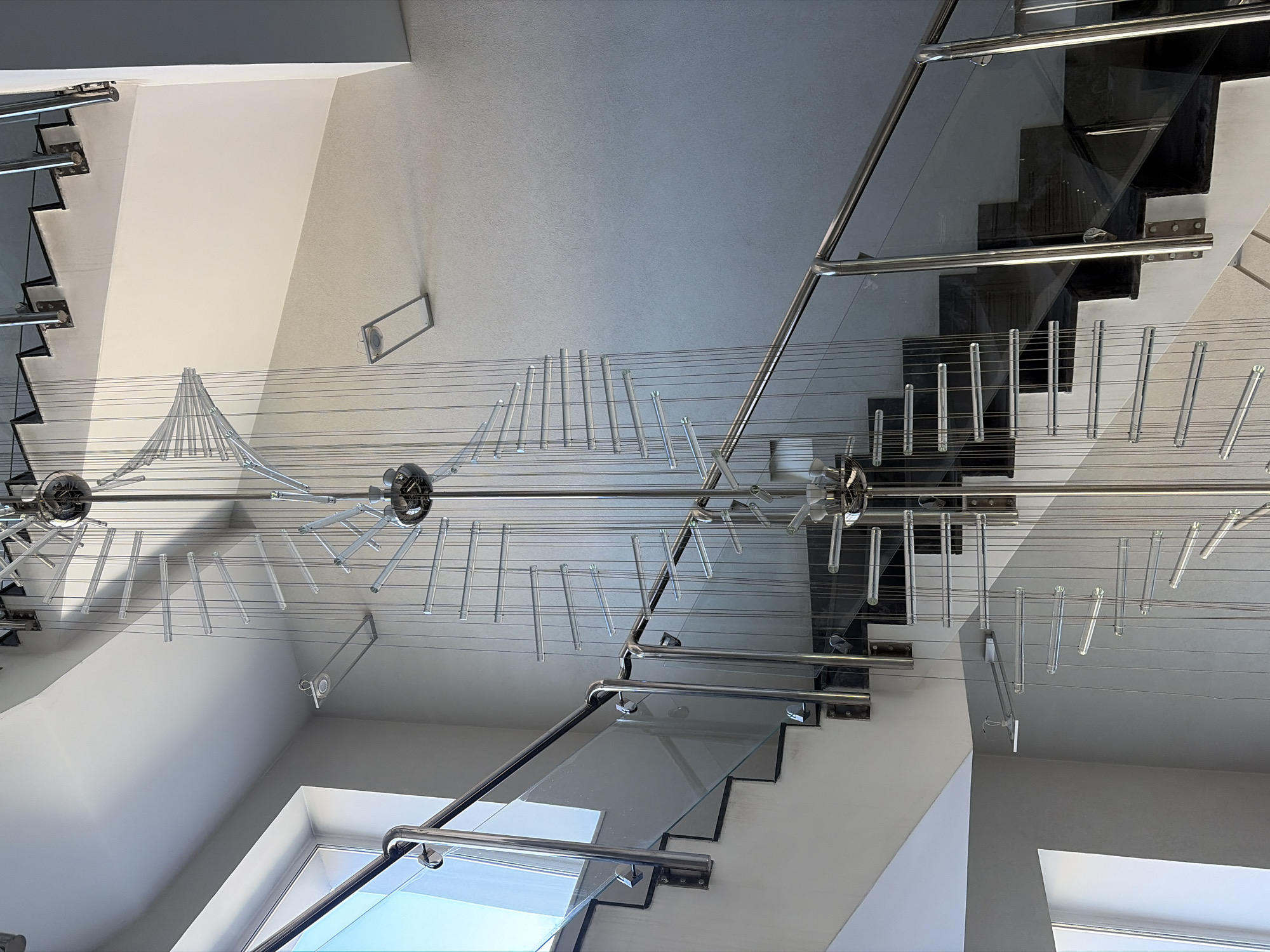 The image shows a modern interior, possibly of a building in Saint Petersburg, Russia, although the exact location cannot be determined from the image alone. The scene features a staircase with glass steps and stainless steel handrails. A decorative chandelier hangs from the ceiling, comprised of spherical elements and linear, hanging rods suspended on cables. The walls are painted in neutral tones, with windows allowing natural light to enter. The staircase is positioned within the building's architecture, with the walls defining its boundaries.