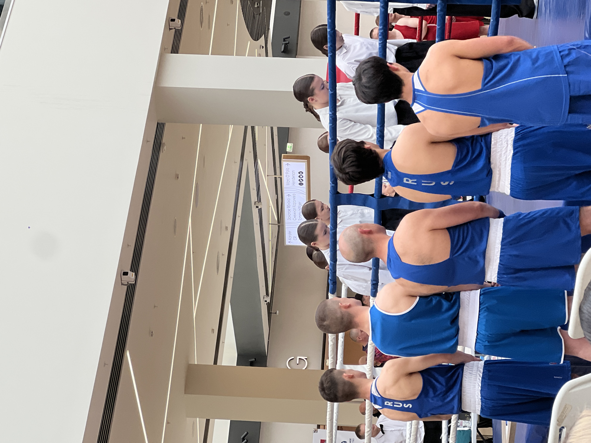 A group of boxers wearing blue uniforms with the letters "RUS" on the back stand near a boxing ring. The setting appears to be a large indoor space, possibly a shopping mall or event venue, with signs directing towards various businesses, including "Kanzler," "Золотое Яблоко," "Снежная Королева," "Marc O'Polo," "Henderson," and "Tommy Hilfiger". The location is in Perm, Russia, based on the Cyrillic text on the sign. Referees in white shirts stand near the boxers.