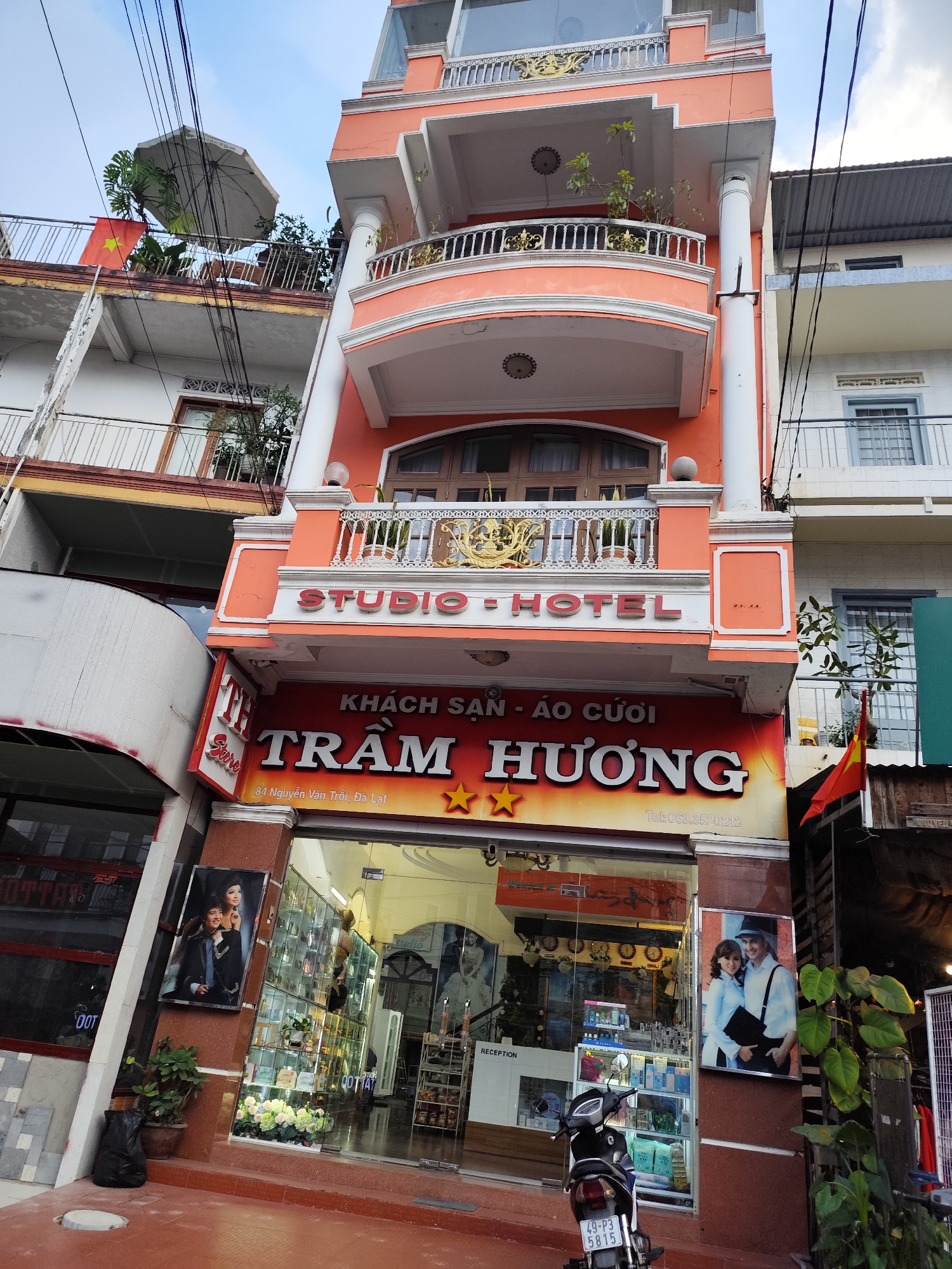 The image shows the "Tram Huong Studio Hotel" located at 84 Nguyen Van Troi in Da Lat, Vietnam. The building is a three-story, peach-colored structure with balconies and decorative railings. A sign with the words "STUDIO - HOTEL" is above a larger sign that says "KHÁCH SẠN - ÁO CƯỚI TRẦM HƯƠNG" with two yellow stars. A motorcycle is parked in front of the entrance. Photos of couples are displayed on either side of the entrance. The interior of the shop, visible through the glass door, shows display cases with various items. A small Vietnamese flag is visible on the right side of the building.