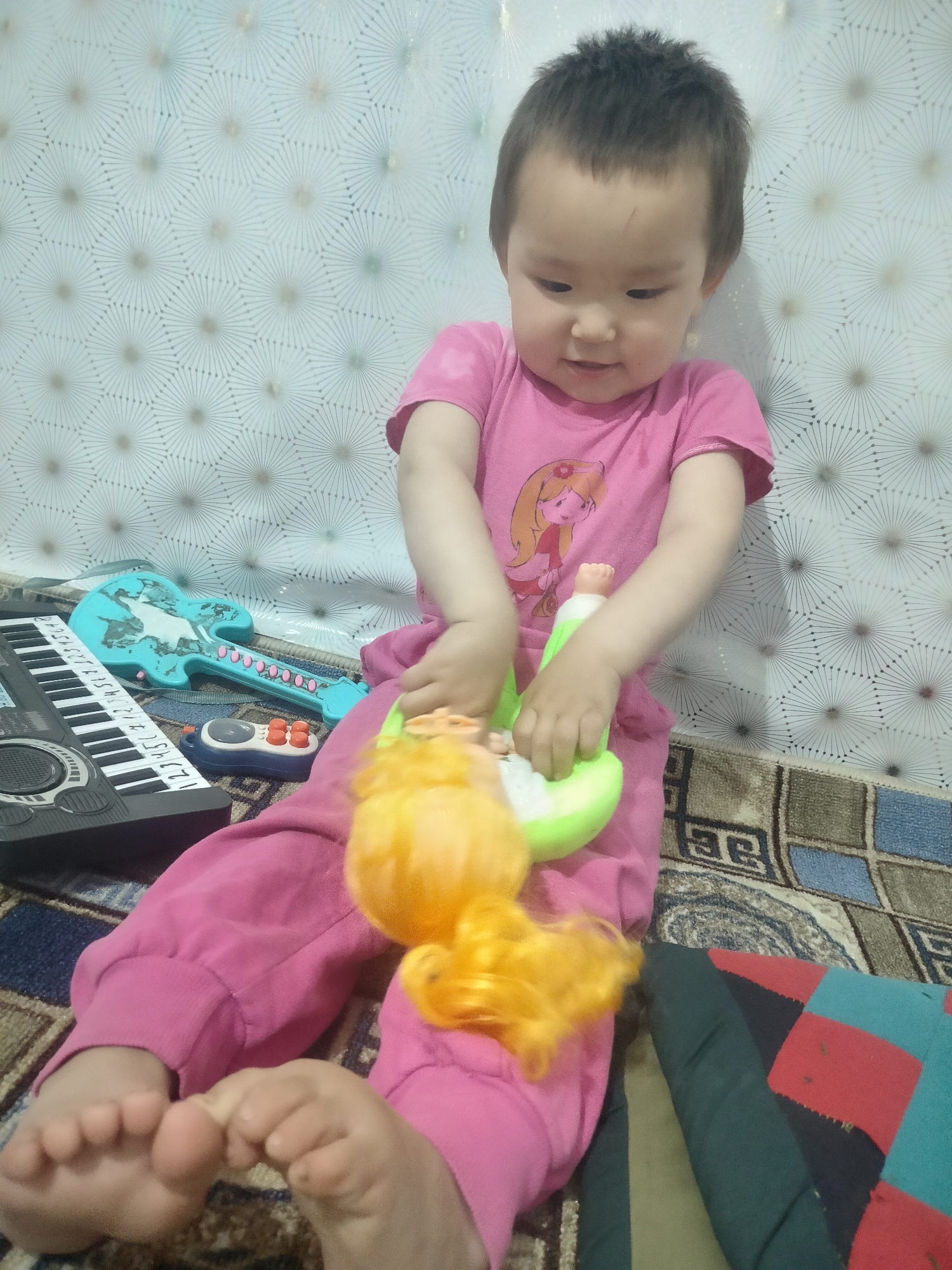 The image shows a young child, appearing to be a toddler, sitting on a patterned rug in what appears to be a room. The child is wearing a pink t-shirt with a cartoon character and pink pants. They are actively playing with a green toy that has a yellow doll-like figure attached to it. In front of the child are two other toys: a blue toy guitar and a black toy keyboard. The background consists of a white wall with a repeating geometric pattern. The lighting suggests an indoor setting, possibly during the daytime. The scene is located in the city of Cholpon, Kyrgyzstan.