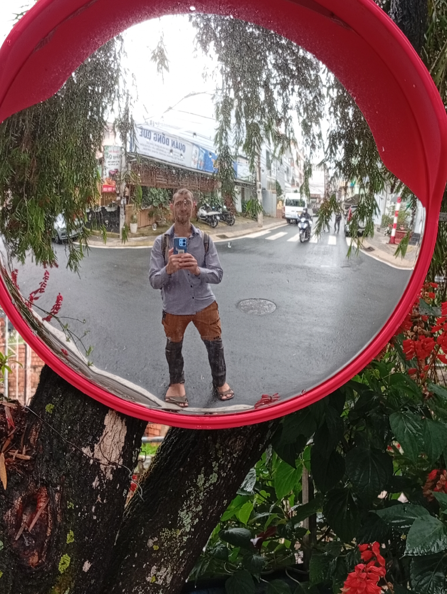 The image shows a round convex mirror with a red frame attached to a tree, reflecting a street scene in what appears to be Da Lat, Vietnam. A man is reflected in the mirror, holding a phone and taking a selfie. He is wearing a light purple button-down shirt, brown shorts, and sandals. He has a backpack on. Behind the man and in the mirror's reflection, there's a street with buildings, motorbikes, and a white van. There are also people walking in the background. The mirror is surrounded by foliage, including a tree trunk and green plants with red flowers. The weather seems wet, as the road surface appears damp. The signage of a nearby business, which appears to be 'QUAN DONG QUE', is visible in the reflection.