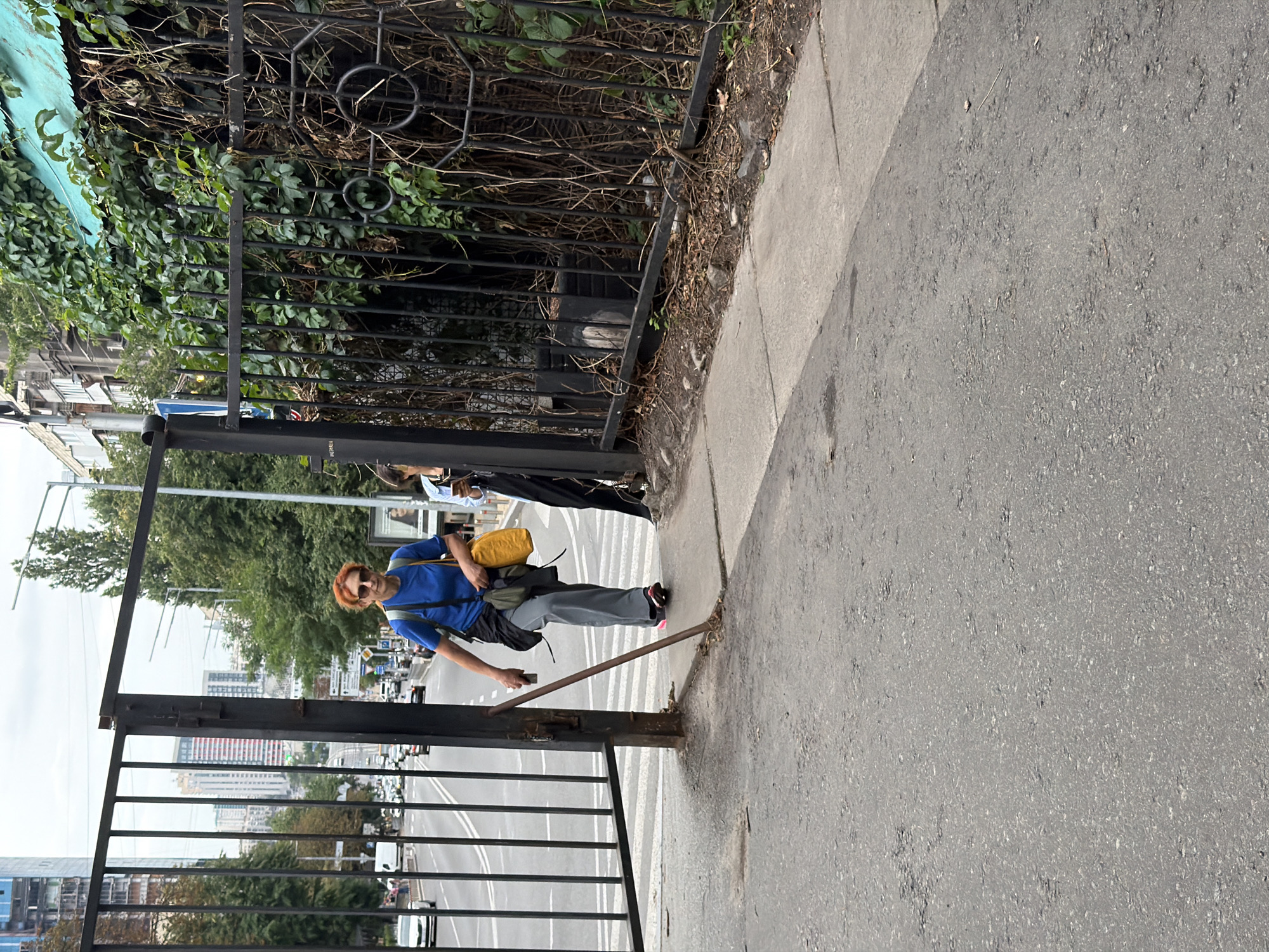 In Kyiv, Ukraine, a woman with short red hair and sunglasses is walking through an open gate on a sidewalk. She is wearing a blue t-shirt, grey pants, and carrying a yellow bag over her shoulder. Another person, partially visible, stands behind her. The gate is made of black metal bars, and beyond it, a street with traffic is visible. In the background, buildings and trees line the street, under a cloudy sky. The sidewalk appears to be made of concrete, and the street is paved with asphalt.