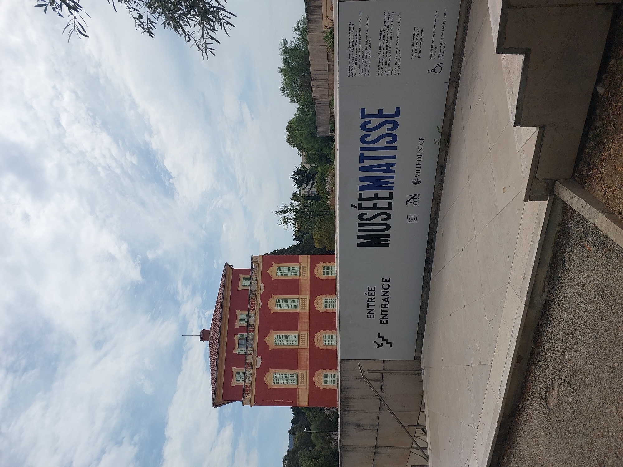 The image depicts the entrance to the Musée Matisse in Nice, France. A large, salmon-colored building with light green shutters and a red tiled roof dominates the background. In the foreground, a large white sign displays the museum's name, "MUSÉEMATISSE," in large blue letters, along with "ENTRÉE ENTRANCE" in smaller black text and an arrow pointing left. Below the name, the logo for "VILLE DE NICE" is visible. To the right of the main sign, there is smaller text providing information about the museum's operating hours and contact details. The ground in front of the sign is paved with large stone tiles, and a gravel path runs along the bottom of the image. Trees and vegetation are visible around the building and to the sides. The sky is partly cloudy. There are no people visible in the image.