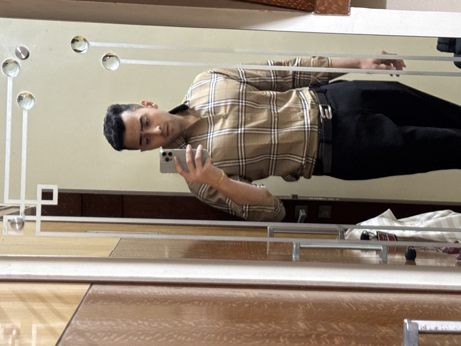 A male individual is depicted taking a mirror selfie indoors. The subject is a young adult male with dark, short, styled hair and clean-shaven facial features, looking directly at the smartphone held in his right hand. He is wearing a long-sleeved, collared plaid shirt in shades of brown, beige, and white, with the sleeves rolled up to the forearms. A thin gold-colored bracelet is visible on his right wrist. He is also wearing black trousers with a black belt and a silver buckle.
The reflection captures him from the waist up. The mirror itself features a decorative border with etched geometric patterns and embedded circular, reflective embellishments. To the left of the mirror is a wooden panel or door with vertical striations. Reflected behind the subject, a dark wooden door with a handle and a light-colored garment, possibly a striped shirt, hanging behind it are visible. A keyring with a red charm hangs near the door handle. The immediate environment suggests an interior space, likely a private residence or a hotel room. While the prompt specifies the city of Kolkata, India, the image itself does not contain any visual cues to ascertain the precise street or building location within the city.