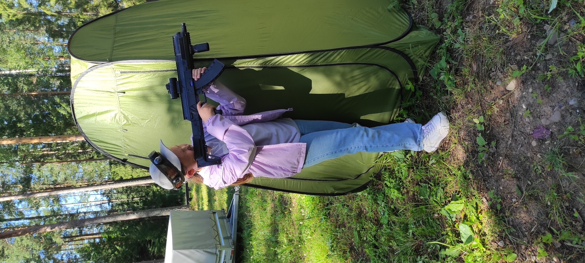 A young girl is holding a toy rifle in front of a green tent in a wooded area. She is wearing a light purple jacket, a gray t-shirt, blue jeans, and white sneakers. She also has a cap with goggles on her head. The tent is open, and it appears to be a portable changing room or shelter. The ground is covered in grass and dirt, and there are trees in the background. A white trailer is visible to the left of the tent. The lighting suggests it is daytime, possibly sunny with some shade. There is no indication of the city of Noviy, Russia, or any urban elements present in the image.