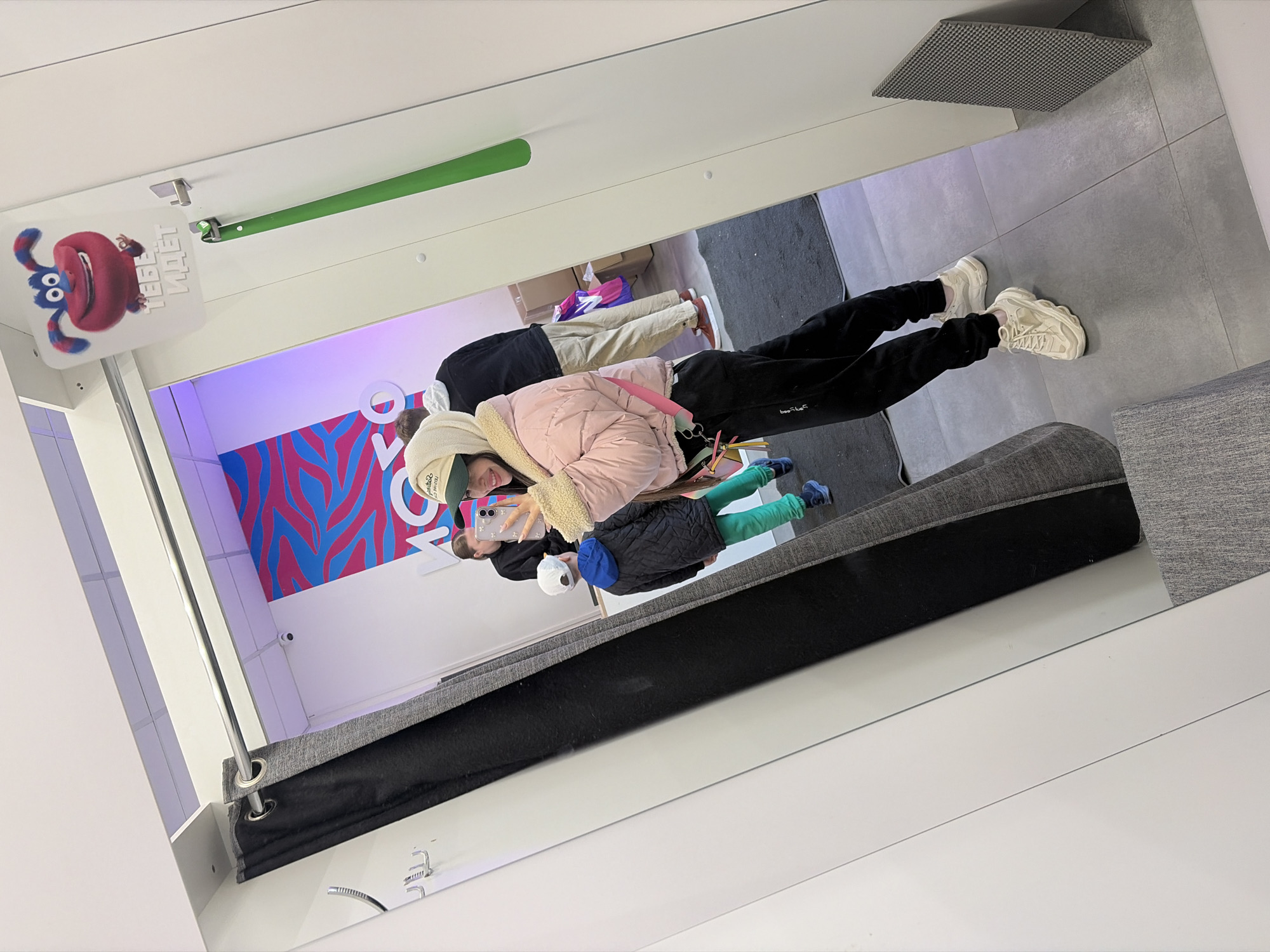 The image depicts a group of people inside a mirror room. A woman in a pink jacket is taking a picture in a mirror. The room has a colorful wall design and a cartoon sign. The location is identified as Saint Petersburg, Russia, based on the sign's text in Russian. The mirror is capturing the reflection of a grey sofa with a black cover.