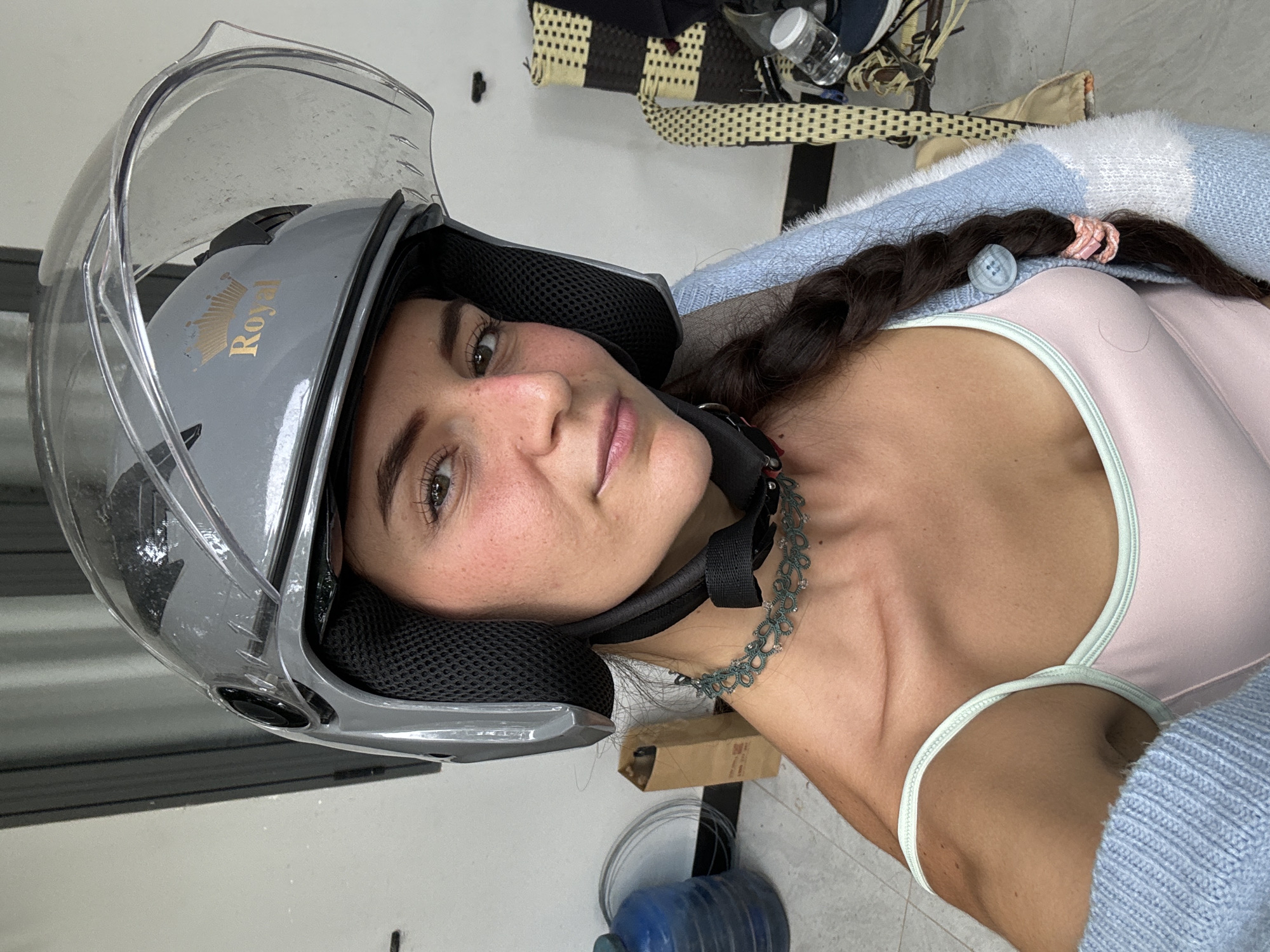A woman is wearing a silver motorcycle helmet and is lying on her side indoors in what appears to be a location in Mũi Né, Vietnam. The helmet has the word "Royal" printed on the side in gold font. The woman has dark hair in a braid, fair skin with freckles, and is wearing a light pink top with a light blue sweater over it. She is also wearing a necklace. To the upper right of the image, there are various objects, including a water bottle and what appears to be a bag. The background includes a white wall and floor.
