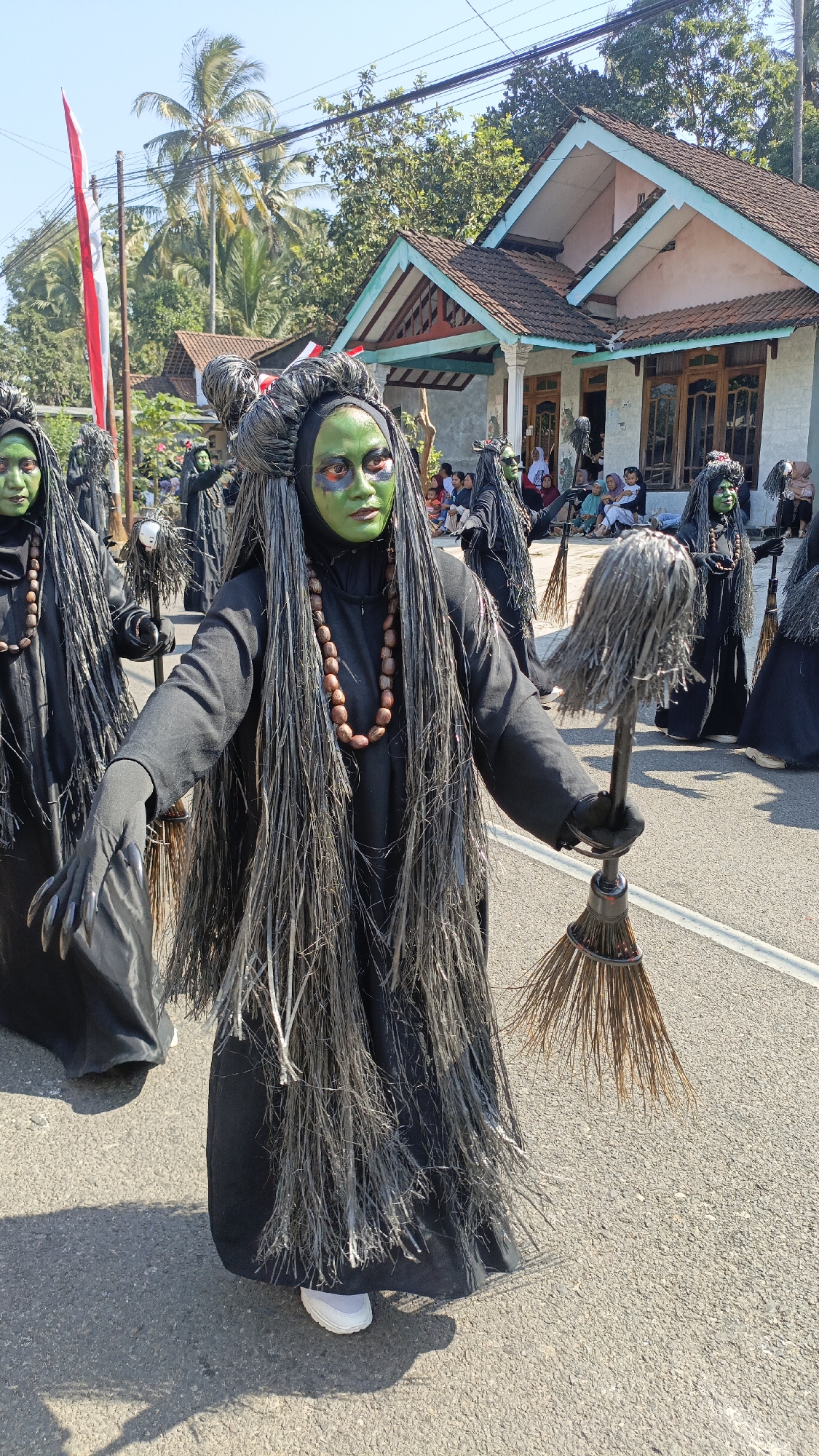In Krajan Lor, Indonesia, a parade is in progress. The foreground is dominated by a person in a black costume with a green face, long black hair, and a necklace of brown beads. This person holds a broom made of straw and what appears to be a mop head. Behind them, other individuals in similar costumes are visible, also holding brooms. In the background, spectators are seated on the side of the road, watching the parade. Houses line the street, with a red and white flag on a pole to the left. Palm trees and other foliage are present, indicating a tropical environment. The sky is clear and blue, suggesting a sunny day.