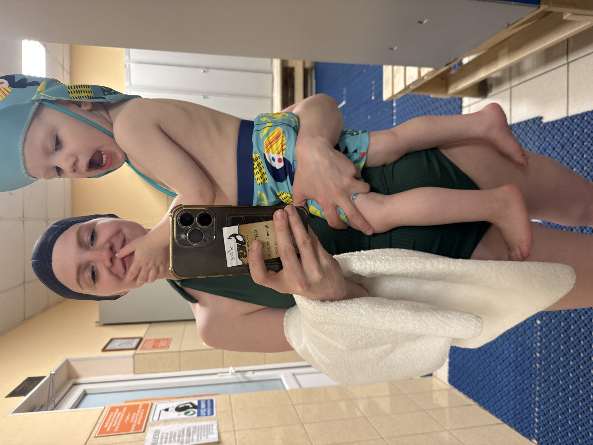 A young woman in a dark green swimsuit and swim cap, holding a toddler in her arms, takes a mirror selfie in what appears to be a locker room, possibly at a swimming pool in Saint Petersburg, Russia. The woman is wearing a black swim cap and has her hand to her mouth. The toddler is wearing a light blue sun hat with a yellow and black pattern and blue swim trunks with a similar pattern. The background shows tiled walls, a door, lockers, and blue mats on the floor. A white towel is draped over the woman's arm.