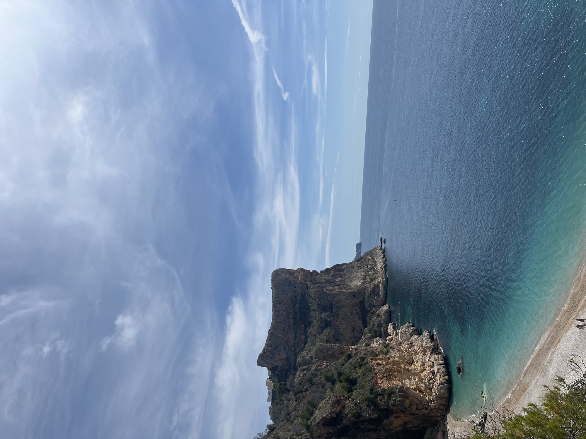 The image shows a coastal scene in el Poble Nou de Benitatxell / Benitachell, Spain. A large, rocky headland juts out into the blue-green sea. The cliff face displays varying shades of brown and gray rock formations, interspersed with patches of green vegetation. Below the headland, a narrow beach curves along the shoreline. The sky is a mix of blue and white, with wispy clouds streaking across it. In the distance, the water extends towards the horizon. The orientation is vertical, suggesting the photo was taken with a mobile device.