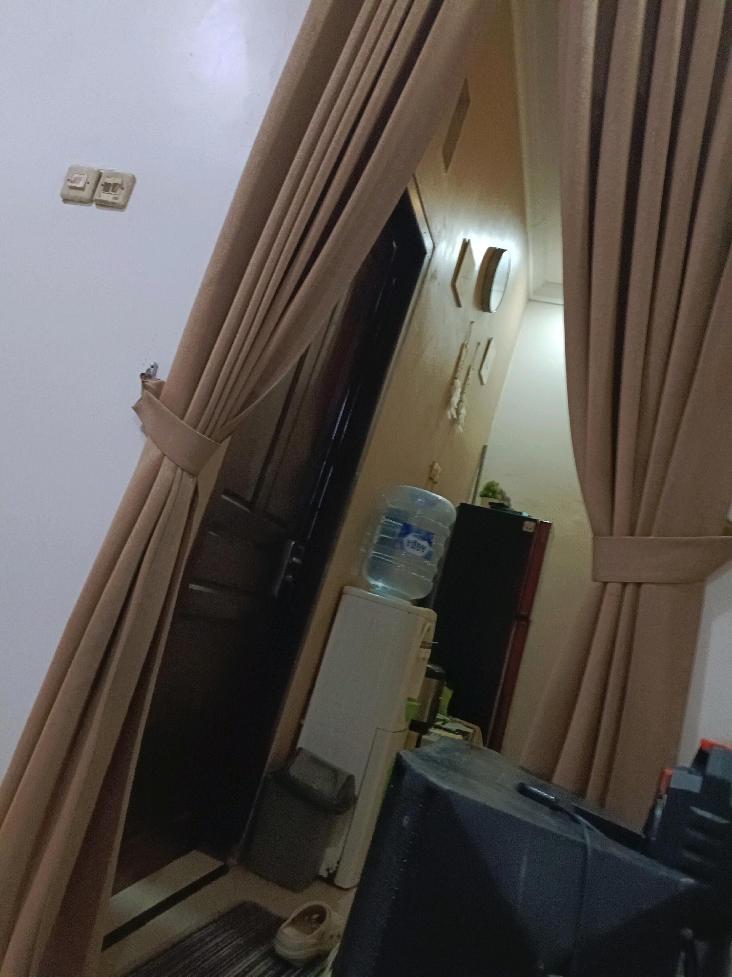 The image depicts an interior scene, possibly a residential hallway or room, with a prominent view of brown curtains drawn to one side. To the left, on a white wall, are two electrical outlets. Further into the scene, a dark wooden door is visible, partially obscured by the curtains. Next to the door, on a white appliance, sits a large bottle of water. Behind the water bottle, a dark refrigerator is present. On the floor in front of the door, a small trash can and a pair of shoes are placed. The overall lighting suggests an indoor environment, possibly with ambient light from unseen sources. The location is identified as Rancang Kencono, Indonesia.