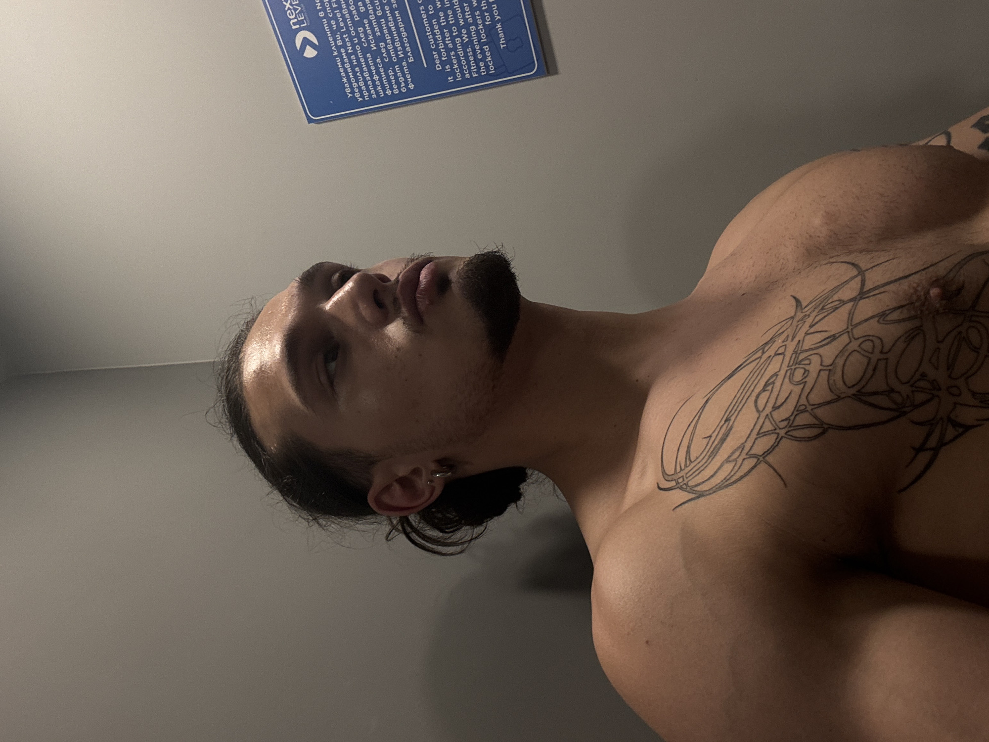 A young adult male with dark hair pulled back in a bun and a light beard is looking to the right in a medium shot. He is shirtless, revealing a large tattoo on his chest. He has a hoop earring in his left ear. The background is a plain gray wall with a blue sign on the right side. The sign contains text in both Bulgarian and English. The English text reads: "Dear customers. It is forbidden to use lockers after the working hours of the fitness. We would like to inform you that according to the rules, the lockers will be emptied every evening after closing time. Thank you for your understanding." The image appears to have been taken in a locker room or gym facility.