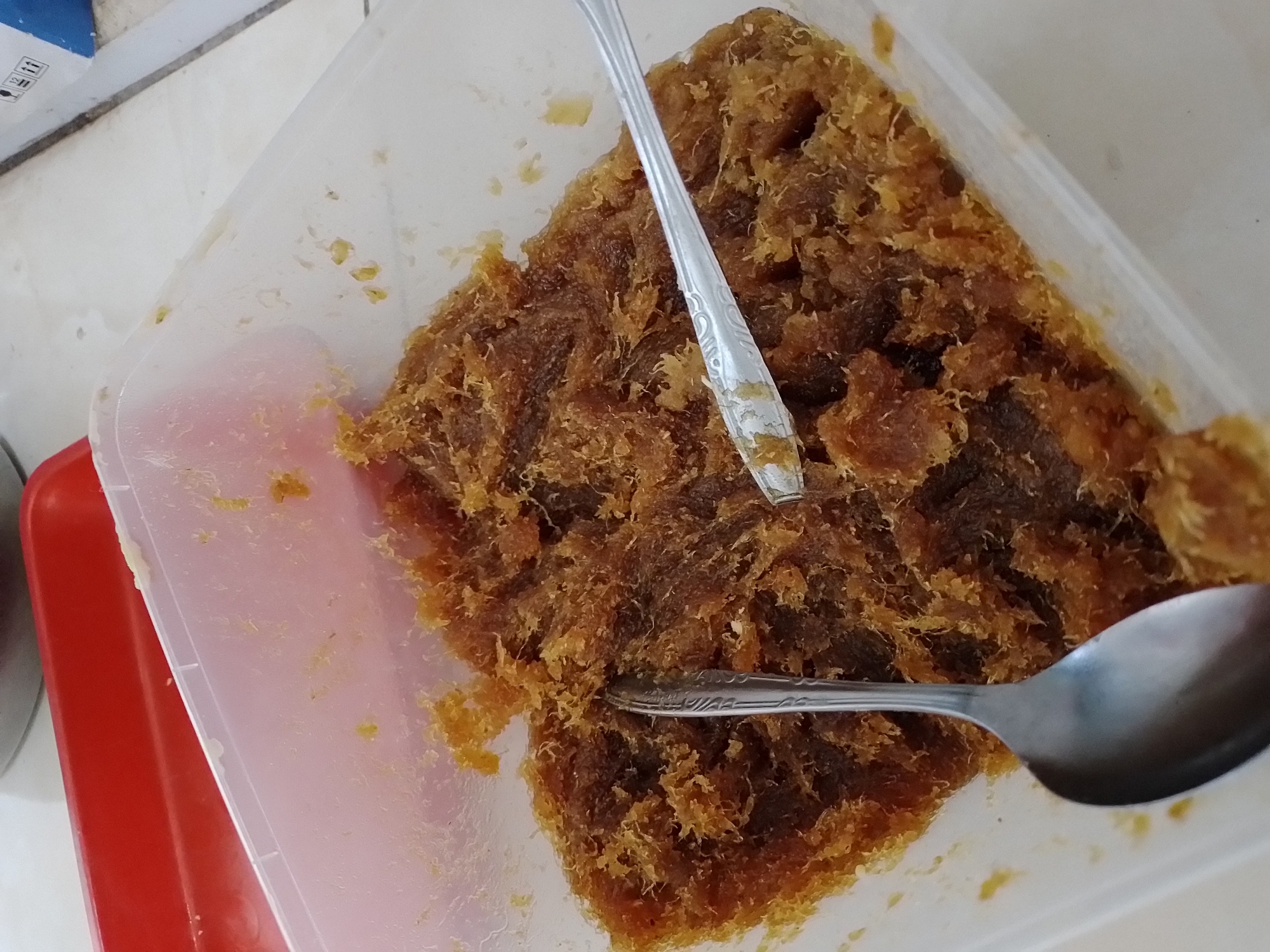 A close-up, overhead shot shows a clear plastic container filled with a light brown, shredded food item. Two metal spoons are partially submerged in the food. The spoon on the left is a large tablespoon, its bowl completely immersed. The spoon on the right is a smaller, flatter spoon, with its edge resting on top of the shredded food. The food itself appears to be made of fine strands, with a slightly glistening texture. The background includes a red rectangular tray placed behind and to the left of the plastic container, and a white surface with some light-colored objects, possibly boxes, in the upper right corner. The scene is lit by overhead light, creating subtle shadows. The image was taken in Ngancar, Indonesia.