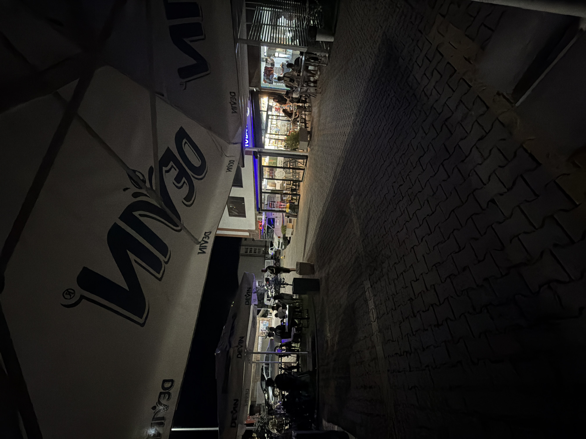 The image depicts an outdoor seating area of a cafe or restaurant at night in Gotse Delchev, Bulgaria. Large umbrellas with the "DEVIN" logo are spread over tables where several people are seated, some interacting and others appearing to be observing their surroundings. A man stands near a parked car, possibly an employee or patron. The ground is paved with interlocking bricks, and building facades with illuminated windows are visible in the background. A white van, possibly a police or emergency vehicle, is parked further down the street, with its lights on. The scene suggests a relatively quiet evening in a commercial area.