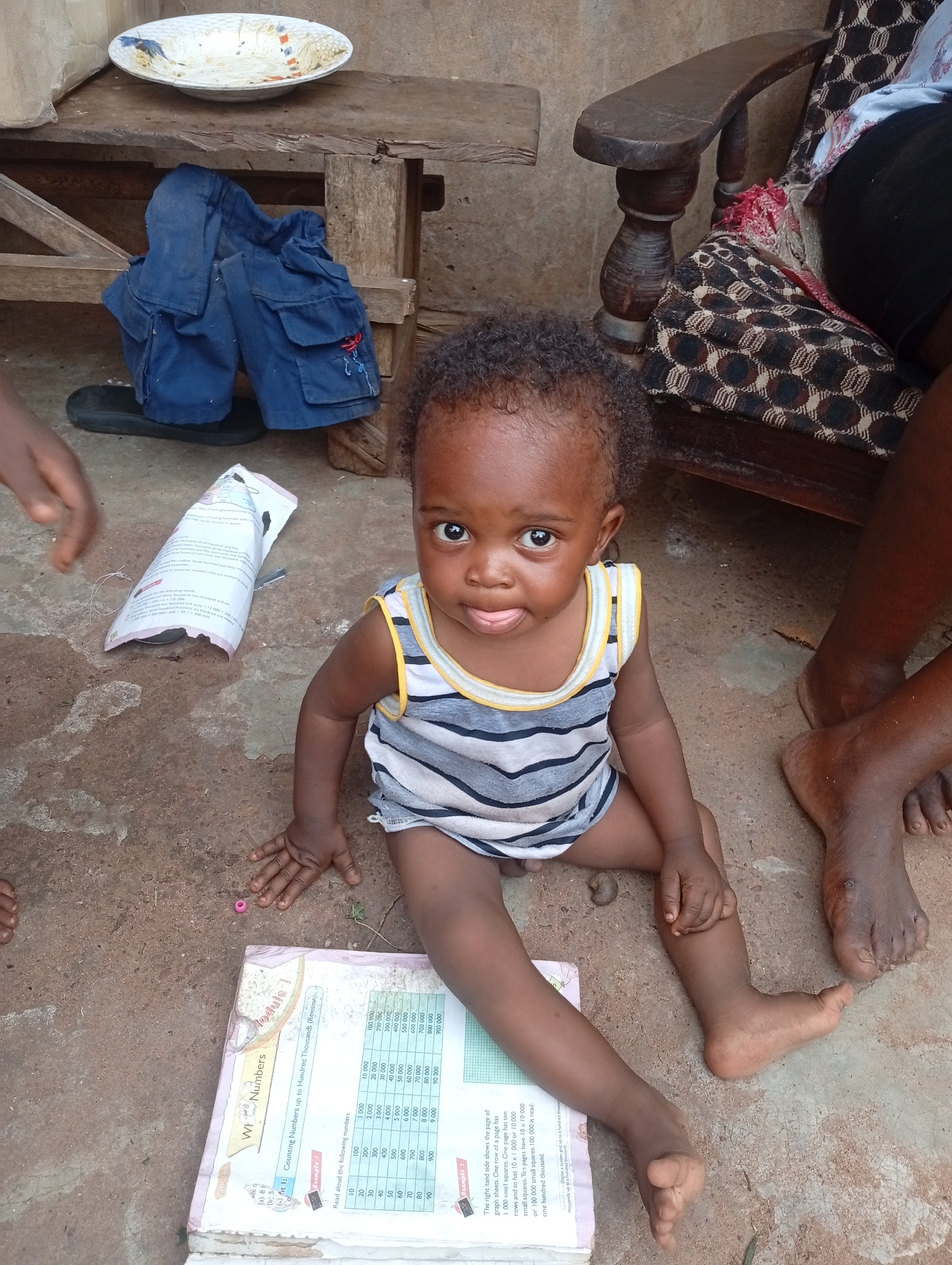 A young child, appearing to be approximately one to two years old, sits on a dirt ground in Adda, Nigeria. The child has dark skin and curly black hair, and is wearing a sleeveless, striped shirt with yellow trim. The child is looking directly at the camera with their tongue slightly out. In front of the child is an open book with text and a grid of numbers. To the left of the child, a hand is visible. To the right, the bare feet of an adult are visible, one resting near the child's leg. In the background, a wooden chair with a patterned cushion is visible, as well as a wooden bench with a blue garment and black sandals placed on it. A white plate with a colorful pattern sits on the bench. The environment appears to be outdoors, with a rustic, unpaved ground.