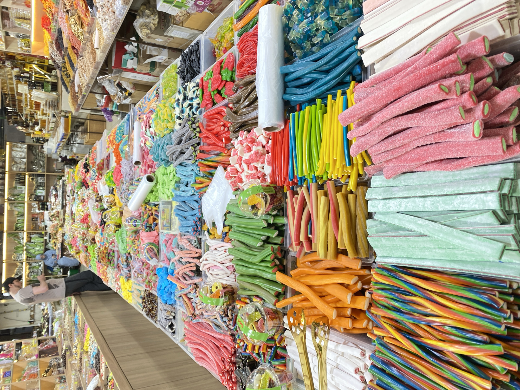The image depicts a vibrant candy store, likely in Makhachkala, Russia, filled with a wide variety of colorful confectionery. Long tables are laden with bulk candies, including gummy worms, licorice sticks, sour belts, and fruit-shaped gummies. The candies are arranged in transparent containers and trays, showcasing their diverse shapes, sizes, and colors, ranging from bright pink and red to green, yellow, blue, and multi-colored options. Some candies are dusted with sugar, while others have a glossy finish. In the background, shelves are stocked with more packaged candies and dried goods. Two individuals are visible in the image: a man in a gray t-shirt and dark cap is standing behind a counter, looking at his phone, and a woman in a light blue headscarf and dress is seated at a table in the distance. The overall environment is that of a bustling marketplace dedicated to sweets.
