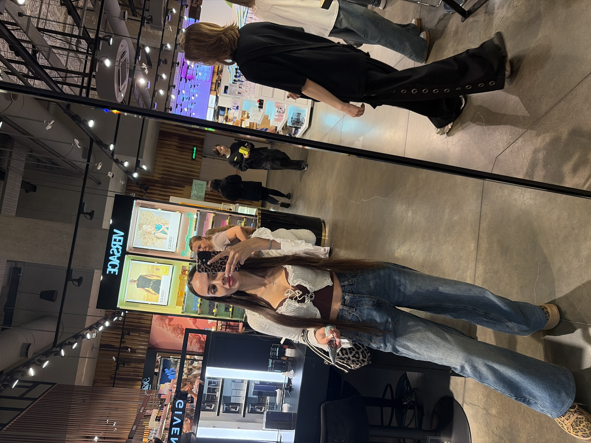 The image was taken in the city of Saint Petersburg, Russia, within a luxury retail store, likely a department store or a high-end boutique, as indicated by the branded displays and illuminated shelves. The store appears to be a beauty or cosmetics retailer, featuring various perfumes and skincare products. The main subject is a woman taking a selfie in a large mirror. She is wearing a white ruffled cardigan over a dark, possibly maroon, crop top, and light-wash wide-leg denim jeans. She has long dark hair and is holding a smartphone with a leopard-print case. Her left hand is holding a small, metallic object.
Reflected in the mirror, several other individuals are visible. To the right, another woman with shoulder-length brown hair is seen from the back, wearing a black t-shirt and black pants with silver eyelets down the side. Further back in the reflection, a person in a black long-sleeved top and black pants walks away, while another individual in a light-colored t-shirt is also visible. The store's interior features modern lighting, including track lights and ceiling fixtures, and a polished concrete floor. The overall ambiance suggests a well-lit, contemporary retail environment.