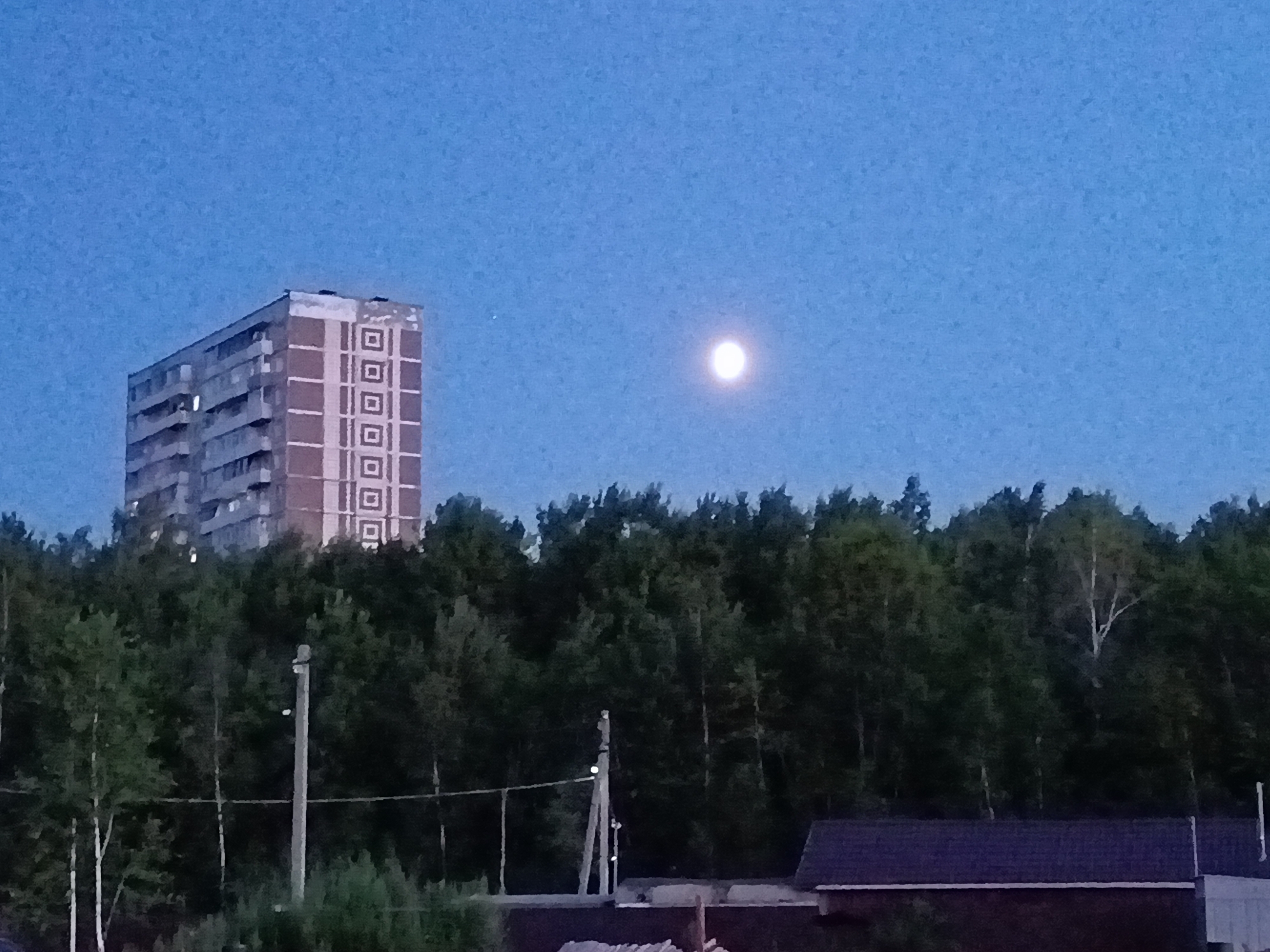 The image depicts a twilight scene in the city of Amursk, Russia. A tall, multi-story brick apartment building with a repetitive square pattern on its facade is visible on the left side of the frame, partially obscured by a dense line of trees. A bright, full moon hangs in the clear blue sky above the trees. The trees, predominantly deciduous, form a thick canopy across the middle ground. In the foreground, a dark-roofed structure, likely a house or a shed, is partially visible on the right. A utility pole with wires is also present in the middle ground. The overall impression is one of a semi-urban or suburban landscape at dusk.