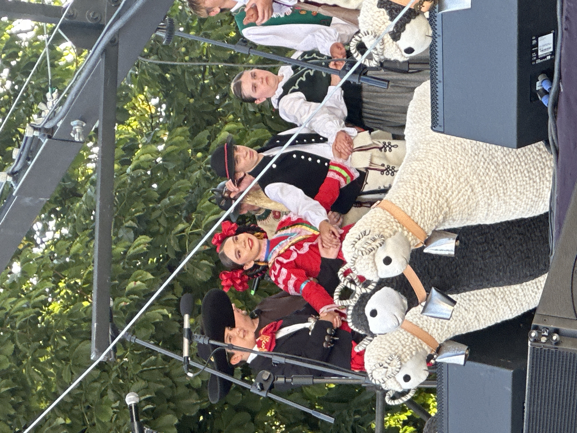 The image depicts a group of young performers on a stage, dressed in traditional folk costumes. In the foreground, three large, knitted sheep figures with bells around their necks are positioned on top of large black speakers. Behind the sheep, from left to right, are several individuals. The first two are children in what appears to be mariachi attire, featuring wide-brimmed hats, jackets with ornate detailing, and sashes. The child on the right is a girl wearing a vibrant red dress with intricate embroidery and red flowers in her hair. Next to her is a boy in a black vest over a white shirt, with decorative stitching on his trousers. To their right, a girl wears a dark vest over a white blouse and a long grey skirt, and another individual to her right is dressed in a white shirt with a green vest. The background consists of lush green foliage, suggesting an outdoor setting. The stage appears to be set up for a performance, with microphones and cables visible. The presence of a stage, speakers, and performers suggests a cultural event or festival. Considering the context of traditional folk costumes and potential performance, this scene is likely taking place during a cultural celebration in Nowy Sącz, Poland, possibly at an outdoor venue or park hosting such events.