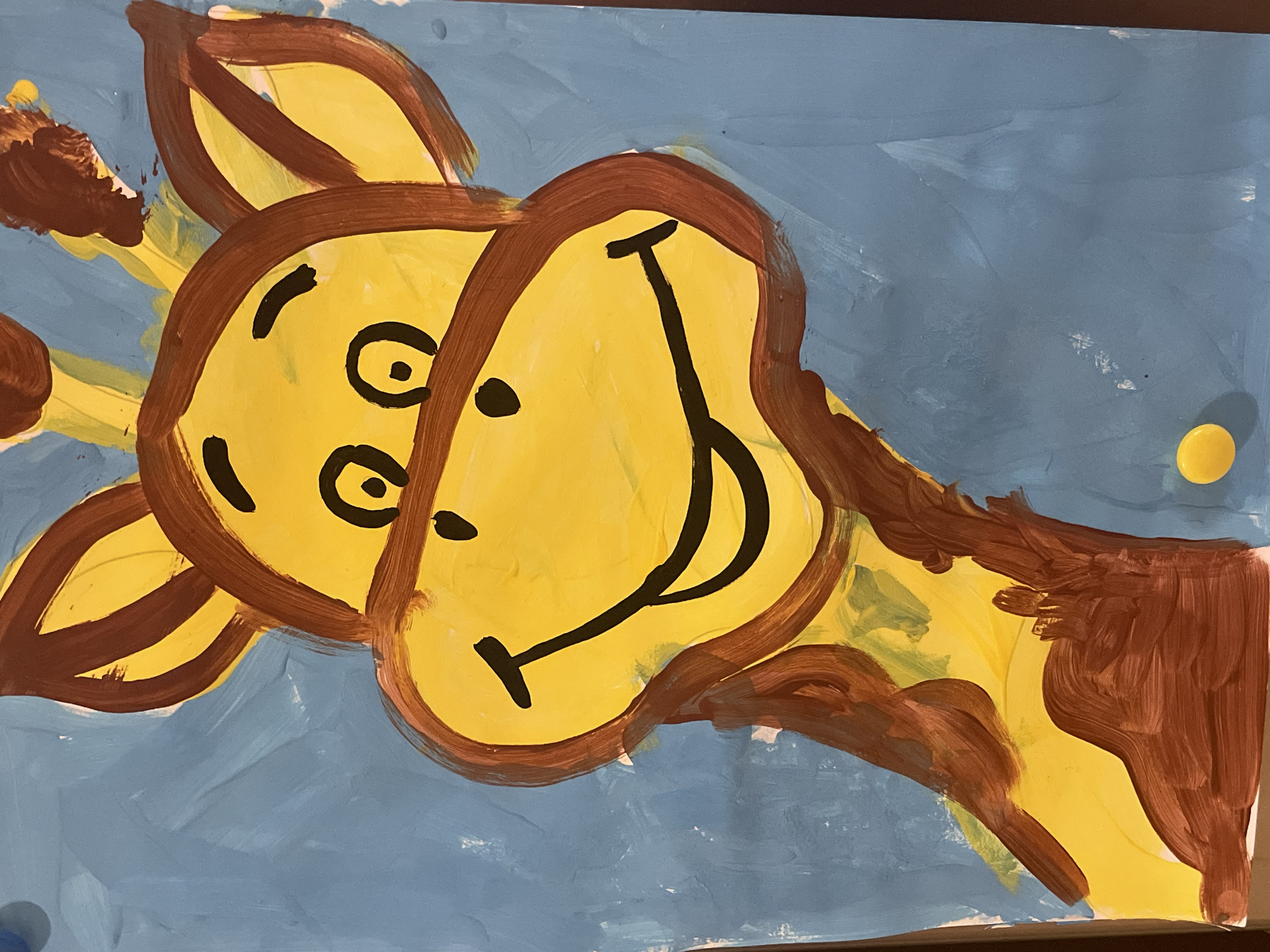 A child's painting of a yellow giraffe with brown spots against a blue background is displayed. The giraffe's head is large and takes up most of the frame. It has a wide, smiling mouth, large, round eyes with black pupils, and two small black dots above its eyes. The giraffe's ears are pointed and outlined in brown. Its neck and upper body are also visible, covered in brown splotches. A small yellow circle, possibly a button or a painted detail, is located in the lower right corner of the image. The painting appears to be on paper and is mounted on a dark surface. The location of this painting is unknown.