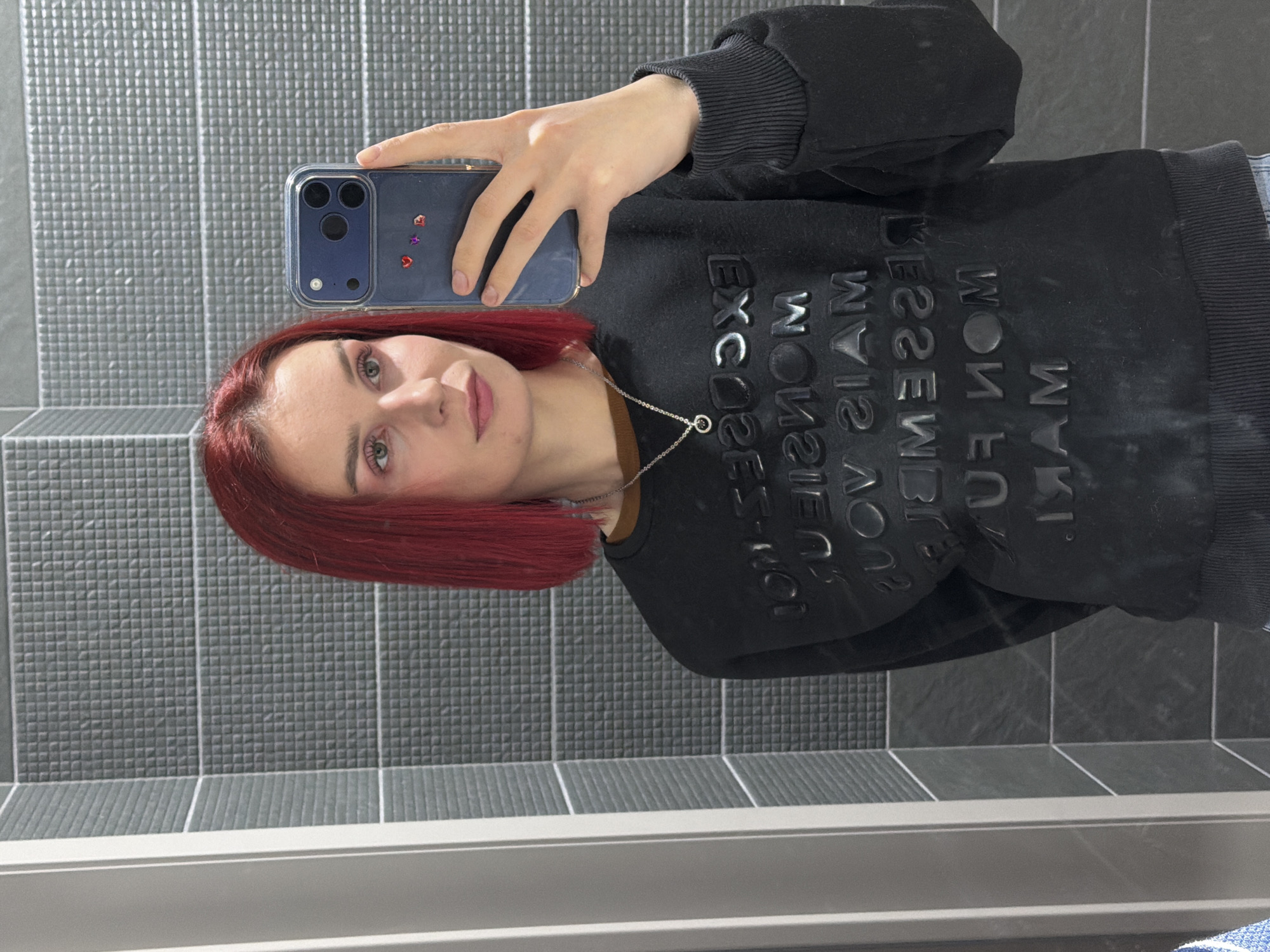 A portrait-oriented mirror selfie features a young woman with distinctive reddish-brown hair and light-colored eyes, looking directly at the camera with a neutral expression. She is wearing a black long-sleeved sweatshirt with prominent, raised text across the chest. Due to the mirror reflection, the text is reversed; when read forward, it appears to say "EXCUSE MY MOUTH" and "EXCUSE MY MANNERS." A silver chain necklace with a small circular pendant is visible around her neck.
She holds a dark blue smartphone with a clear case in her right hand. The phone case features three visible camera lenses and several small, colorful heart-shaped stickers. The background consists of grey tiled walls, with an upper section displaying a fine square grid pattern and a lower section showing broader horizontal lines, separated by a light-colored grout line. A white or light grey ledge runs across the bottom of the frame. The image captures an indoor setting, likely a bathroom or hallway, located in Chaikovsky, Russia.