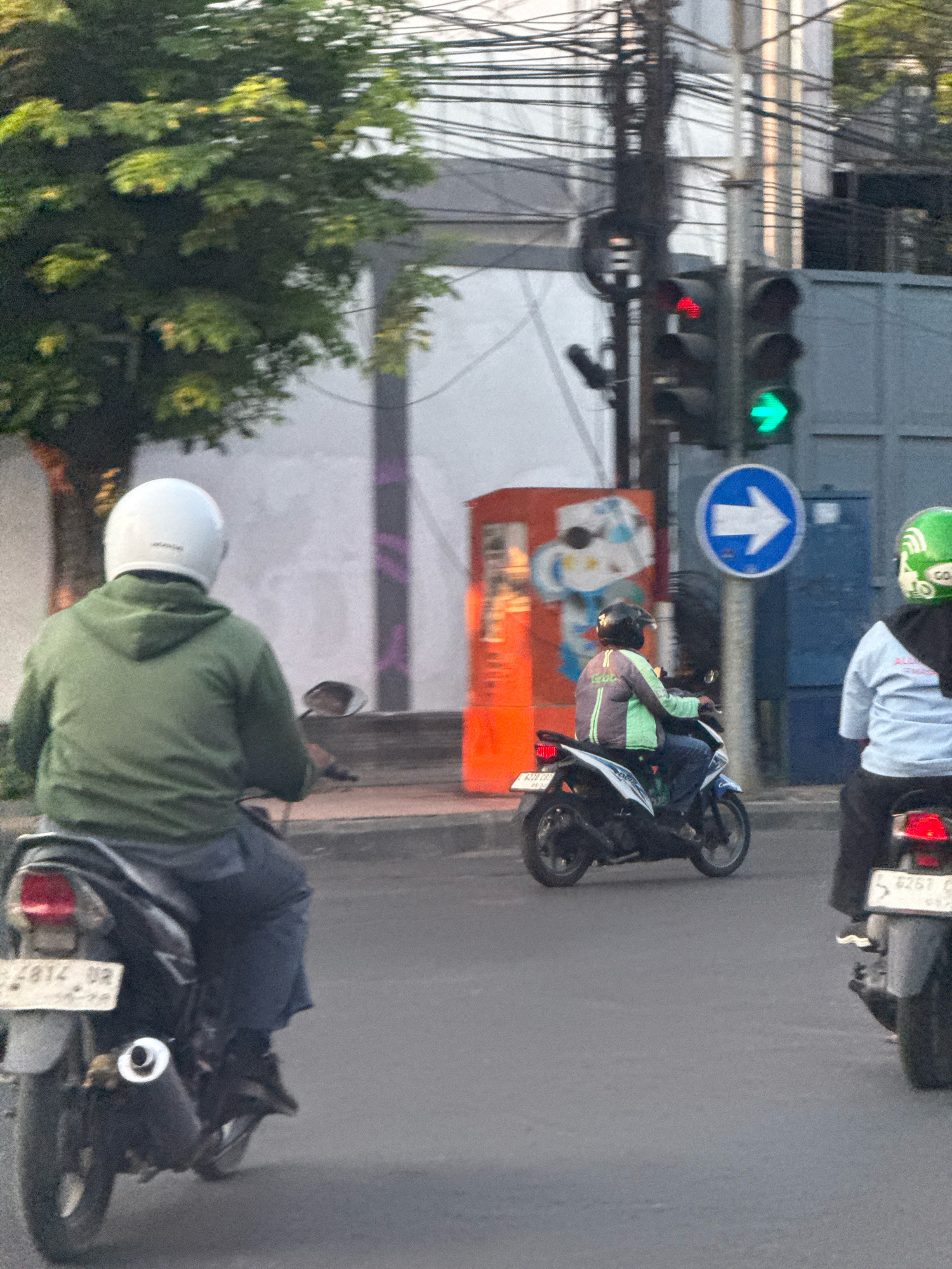 In this medium-range, outdoor, eye-level shot, three motorcyclists are captured on a street in Surabaya, Indonesia, likely during daylight given the ambient lighting. The focus is on the rear of the lead motorcyclist, who is wearing a white helmet and a green hooded jacket, riding a black motorcycle with a visible license plate ending in "02". To the right and slightly behind, another motorcyclist in a green and grey jacket, also wearing a helmet, is on a white and blue motorcycle. Further to the right, a third motorcyclist in a light blue shirt and green helmet is on a black motorcycle. The street is paved, and a sidewalk is visible in the background with a traffic light showing a red left arrow and a green right arrow. There is also a blue circular sign with a white arrow pointing to the right. A tree with green foliage is to the left of the frame, and a vibrantly colored mural is visible on a structure in the background. Electrical wires are prominent above the street.