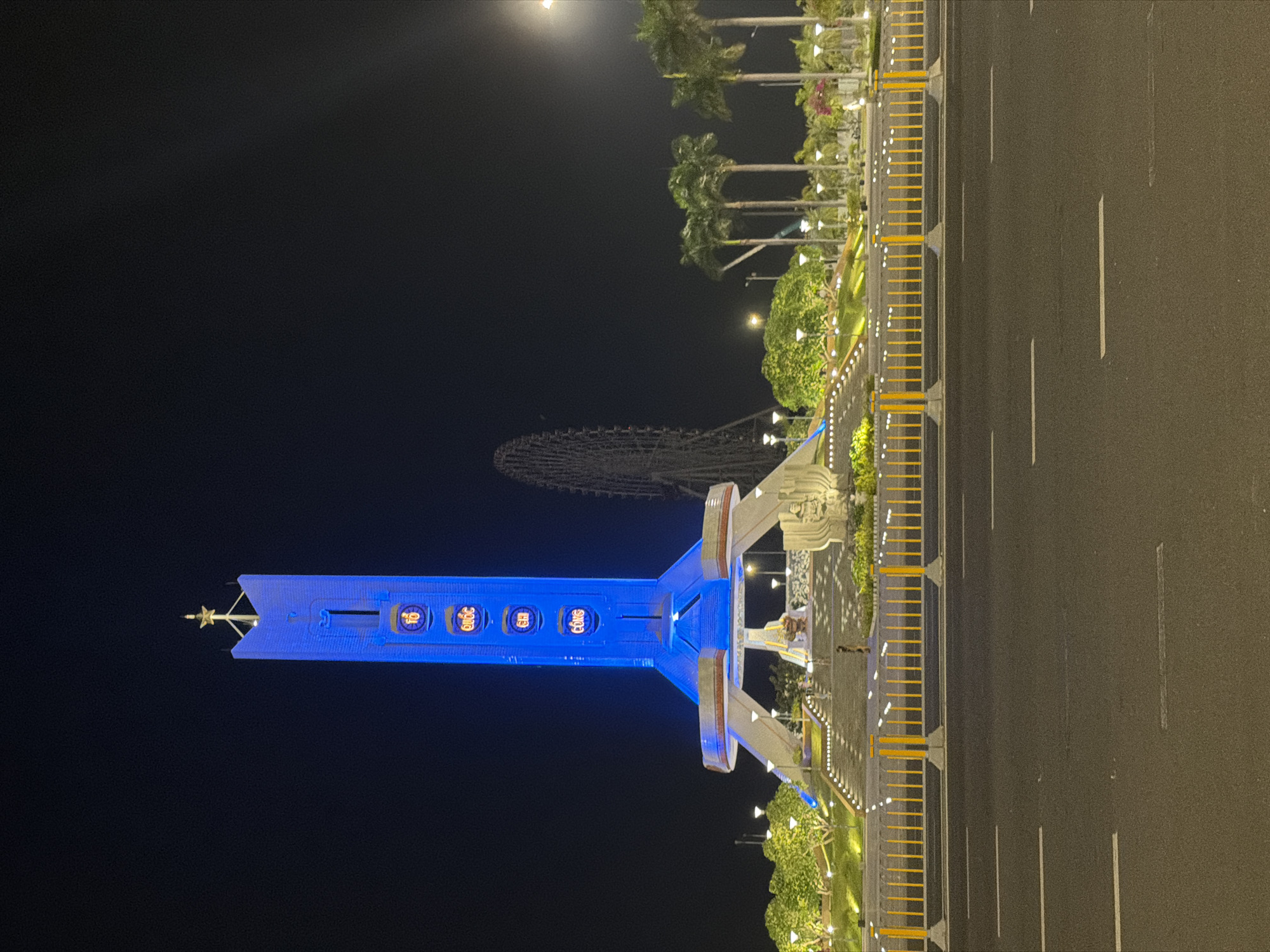 This is a night-time photograph of the Dragon Bridge in Da Nang, Vietnam. The bridge is illuminated with blue and yellow lights, and the surrounding area is also lit up with various lights and palm trees. There are cars driving on the road in front of the bridge. The image appears to be taken from a low angle, looking up at the bridge. The sky is dark and clear, with no visible clouds. There is a ferris wheel in the background.