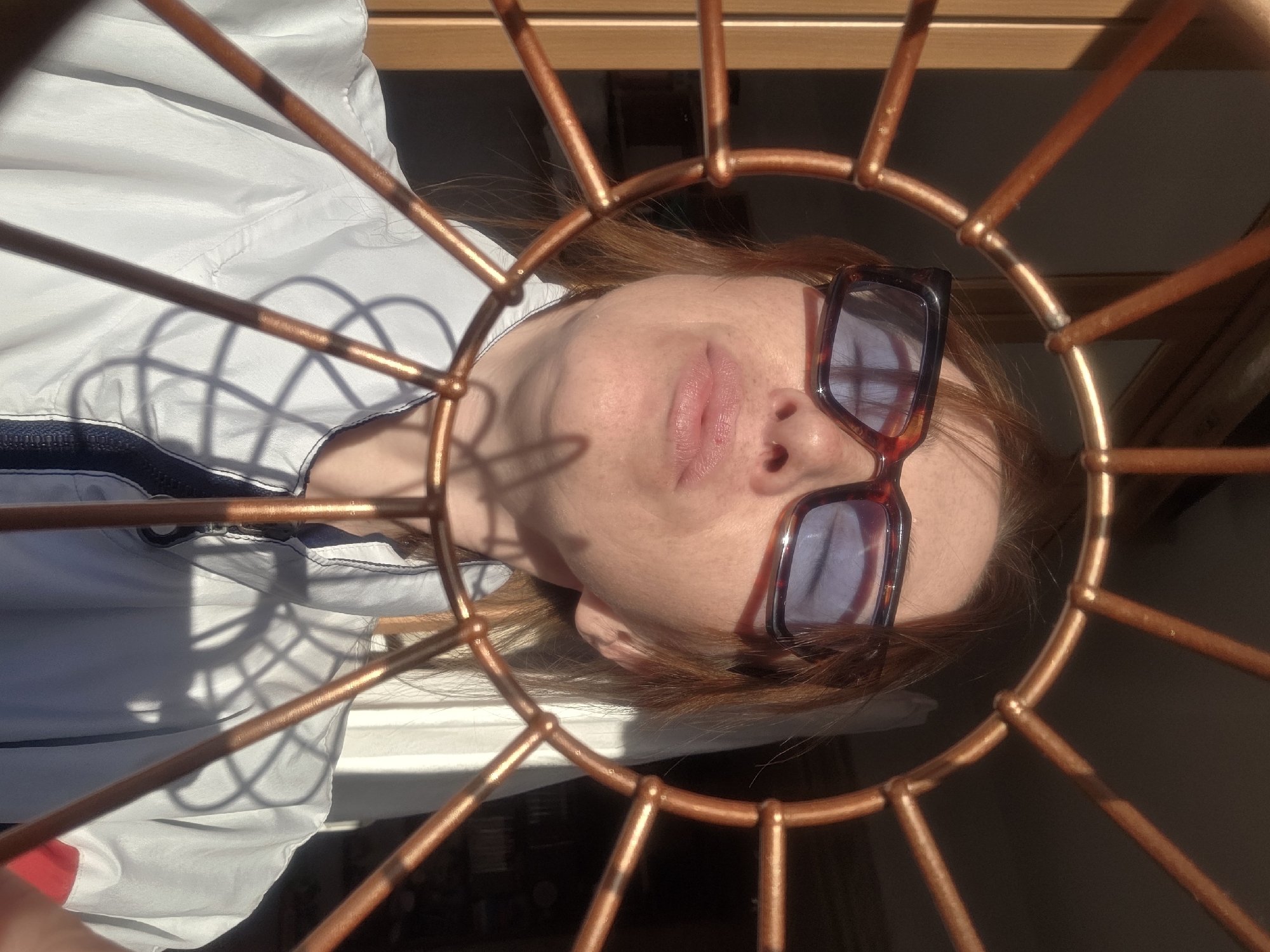 A woman with reddish-brown hair and wearing sunglasses is framed by a circular metal structure with radiating bars. She has fair skin and freckles. She is wearing a white jacket with a blue stripe. The background visible through the metal structure appears to be a light-colored wall or window. The specific location in the city of Chaikovsky, Russia, cannot be determined from the image alone.