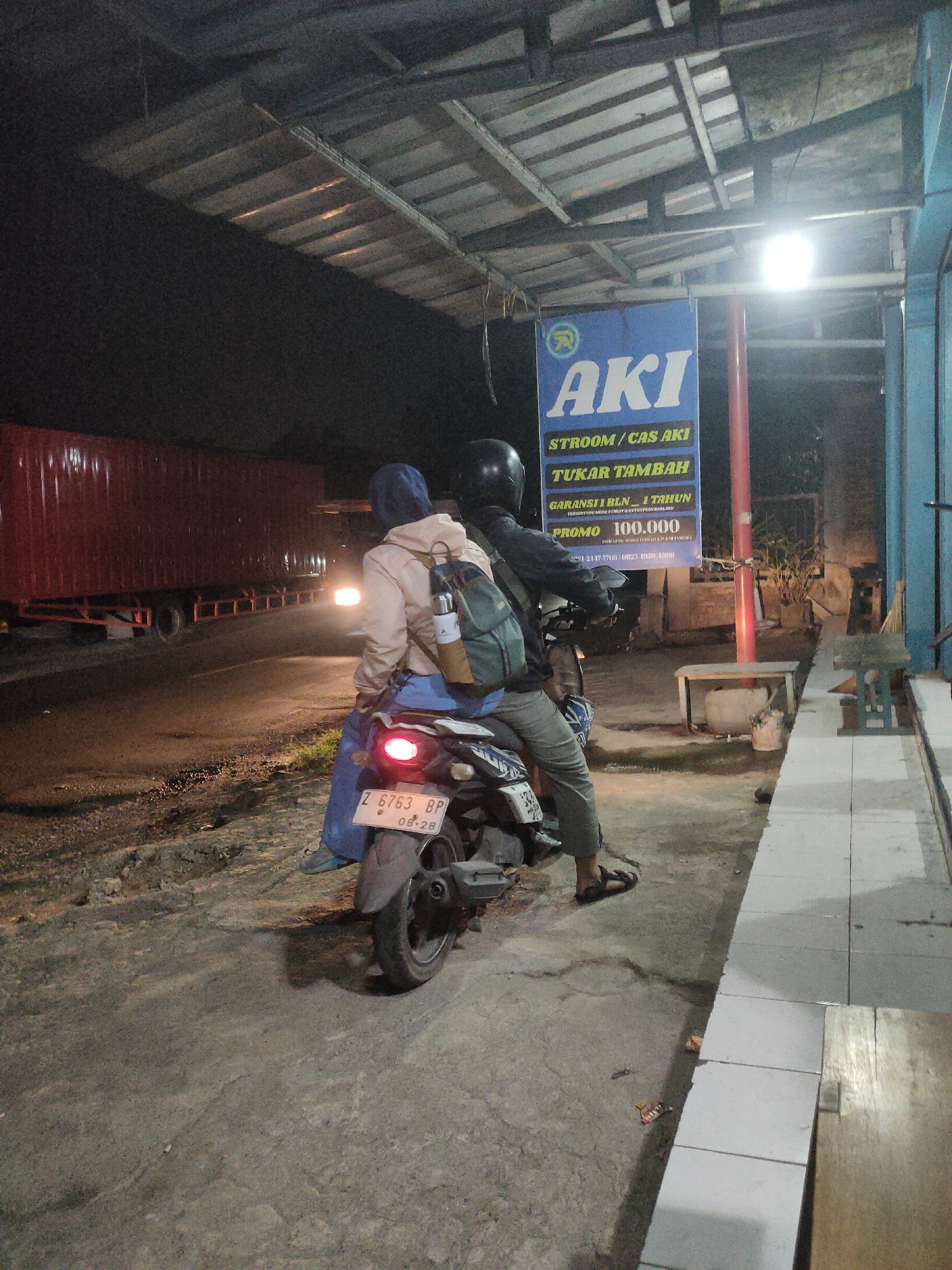 At night, two individuals are seen from behind riding a motorcycle in what appears to be an outdoor area with a covered structure overhead. The motorcycle is parked next to a sidewalk, and a large red truck is visible on the road in the background. A blue sign advertising "AKI STROOM / CAS AKI TUKAR TAMBAH" with promotional details is prominently displayed on a red pole. The ground is asphalt, and there is some debris scattered around. The scene suggests a roadside service area or a commercial establishment in Sumedang, Indonesia, possibly a battery shop.
