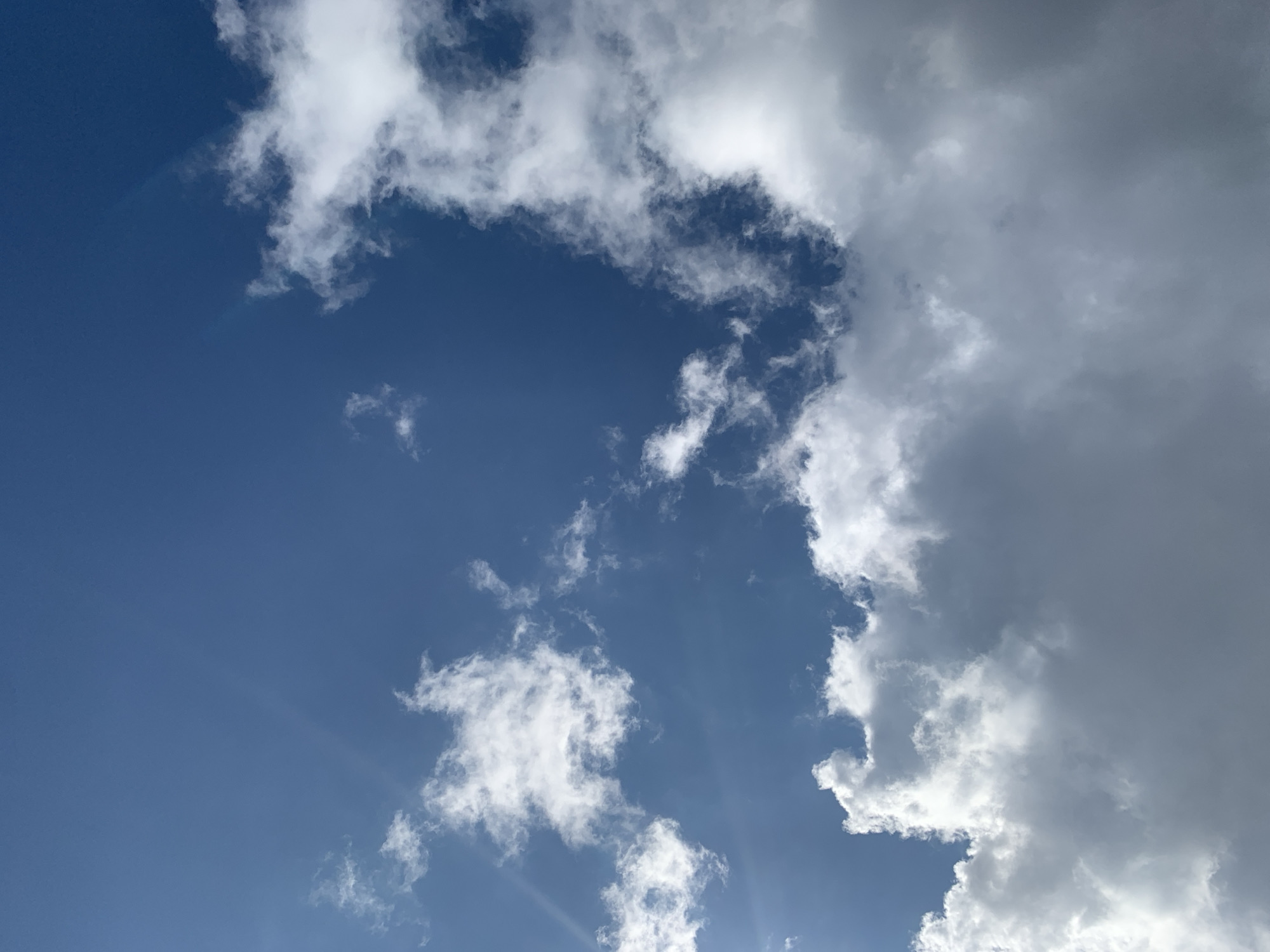 The image is a close-up, ground-level view looking directly upwards at a vibrant blue sky filled with a mix of white and gray clouds. The lighting suggests it is daytime, with bright sunlight filtering through some of the clouds, creating a lens flare effect in the upper left quadrant. There are no people, buildings, or other discernible objects or features visible in the frame, focusing solely on the atmospheric conditions. The scene depicts a typical sky in Lviv, Ukraine, characterized by partly cloudy weather.