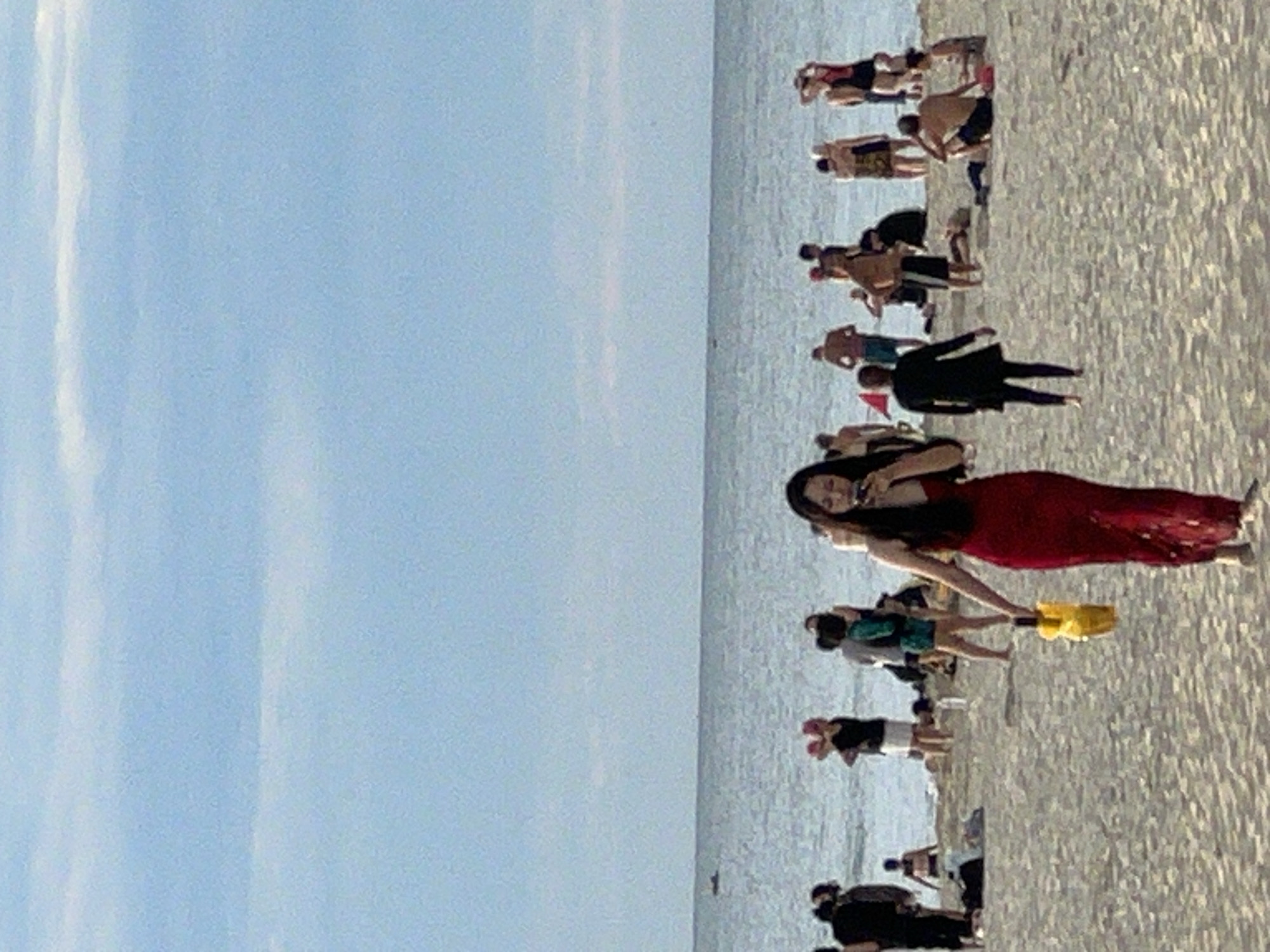 The image depicts a crowded beach scene under a clear blue sky with scattered clouds. In the foreground, a woman in a long, red dress is walking towards the viewer, holding a yellow object. She appears to be looking at her phone. Behind her, numerous people are scattered across the sandy beach, some standing, some sitting, and some in the shallow water. The ocean is visible in the background, with a few small boats in the distance. The overall atmosphere is one of leisure and recreation on a sunny day. The presence of numerous people in swimwear suggests a popular tourist destination. This scene is consistent with a location like My Khe Beach in the city of Đà Nẵng, Vietnam, known for its wide sandy shores and popularity among both locals and tourists.