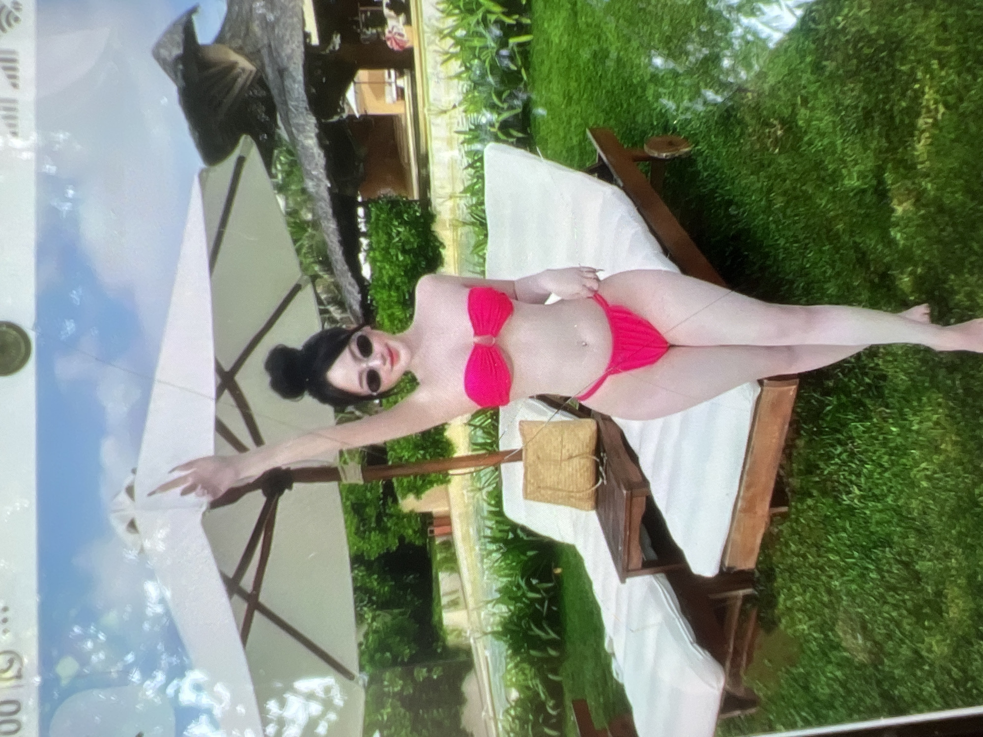 A person in a bright pink bikini is lying on a white lounge chair in an outdoor setting. They are wearing sunglasses and have dark hair pulled back. Their left arm is extended, and their right hand is resting on their bikini bottom. A woven box sits on the lounge chair next to them. The lounge chair is placed on a grassy area, with a large umbrella behind it. In the background, there are trees and a building with a red roof. The image appears to have been taken during the daytime, with bright sunlight illuminating the scene. The overall mood seems relaxed and leisurely, suggesting a vacation or leisure activity. There is no visible text in the image.