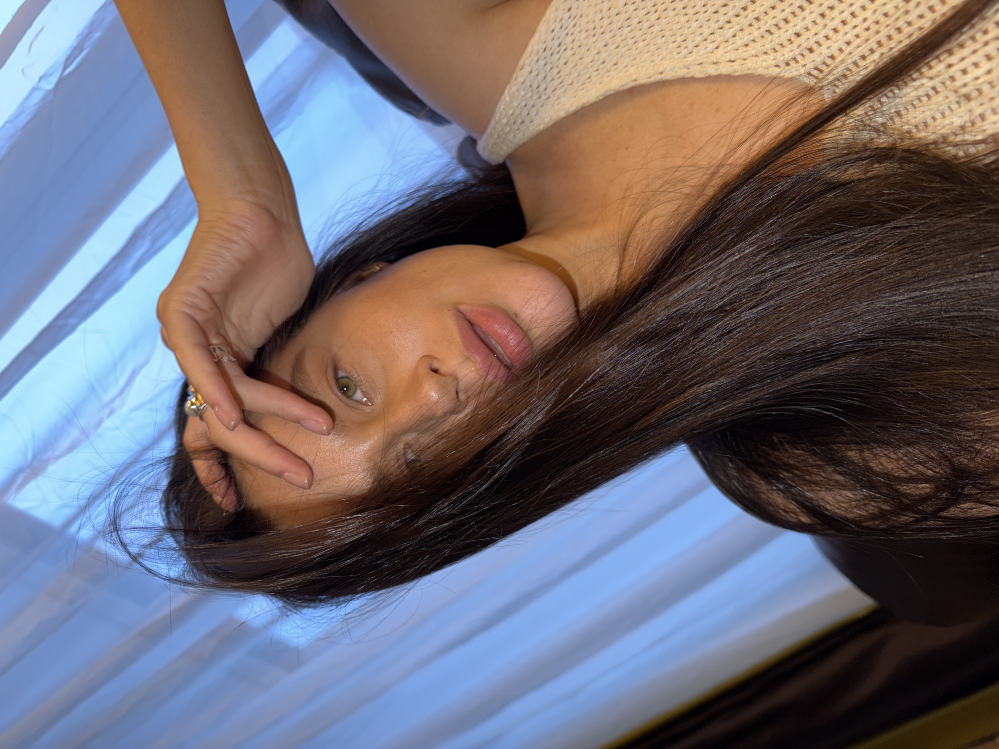 The image, taken in Khimki, Russia, features a young woman lying down or reclining, captured from an overhead perspective and rotated on its side. She is the central subject, with her upper body, face, and long dark hair prominently displayed.
Her gaze is directed towards the viewer, with her green eyes partially visible. Her lips are slightly parted, showing a soft pink tint. Her skin appears smooth, and there's a subtle sheen on her forehead and nose. Her long, dark brown hair spreads out around her head and shoulders, covering part of her face on one side.
She wears a light-colored, possibly cream or beige, knitted garment with a distinct texture, visible on her shoulder and neckline. Her right hand is raised, with fingers gently touching her forehead and temple area, revealing manicured nails with a neutral polish. A gold-toned ring with a clear or light-colored stone is visible on one of her fingers.
The background consists primarily of vertically striped fabric, likely curtains or blinds, in shades of light blue and white, suggesting an indoor setting with a filtered natural light source. A darker, indistinct object or surface, possibly furniture, is present in the lower right corner of the frame. The overall lighting is soft, creating a gentle atmosphere. No text is visible in the image.