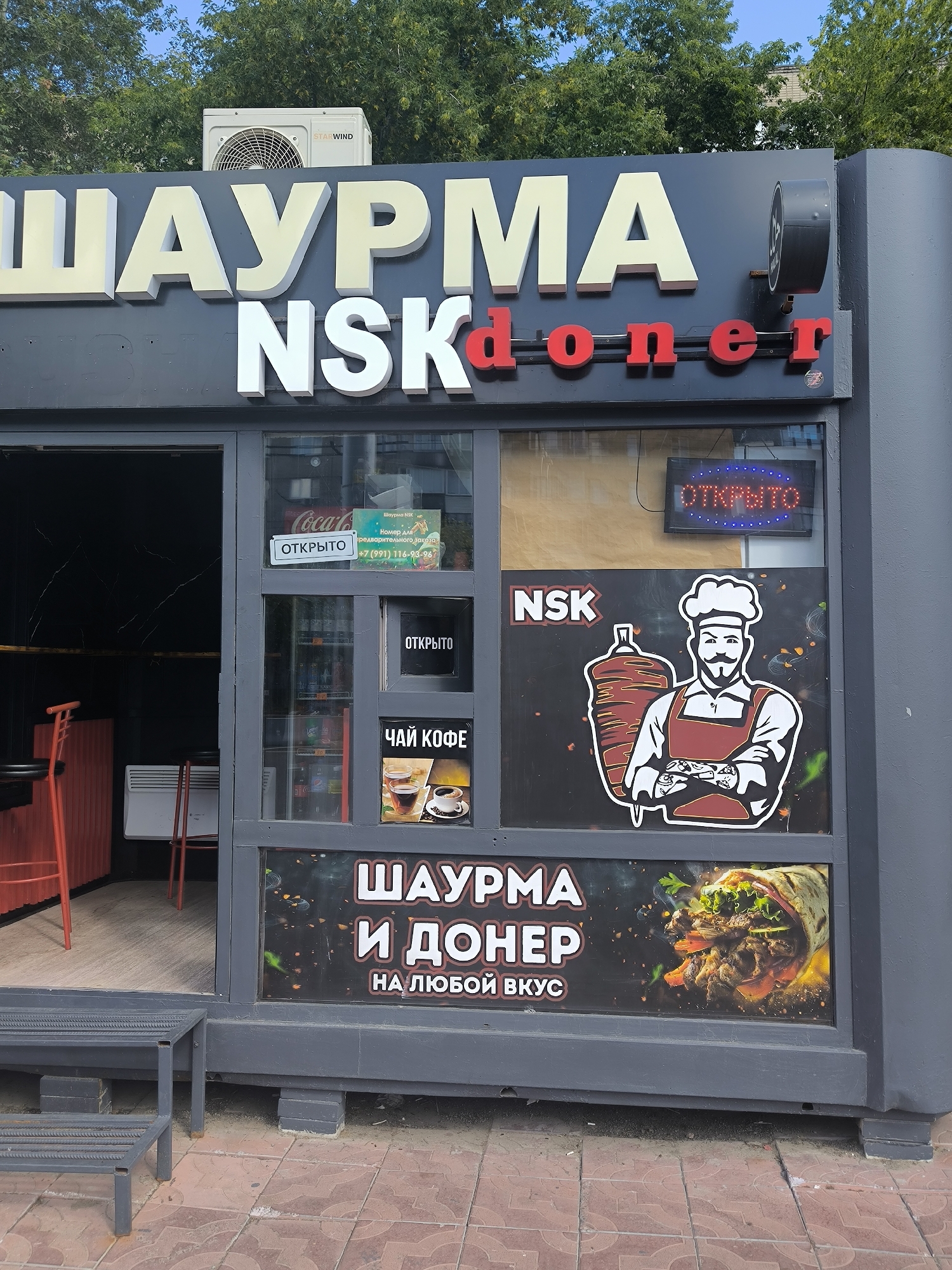 A storefront for a business named "ШАУРМА NSKdoner" is visible. The facade is dark gray with large, three-dimensional white lettering spelling "ШАУРМА" and smaller red lettering spelling "doner" on the upper part of the sign. Below this, in white lettering, is "NSK". A circular black sign with a white emblem is attached to the right side of the facade.
The storefront features large glass windows. On the left window, a red chair and a small table are visible inside. The right window is divided into sections. One section shows a sign with "ОТКРЫТО" (Open) in white text on a blue background. Another section displays an advertisement for "ЧАЙ КОФЕ" (Tea Coffee) with images of coffee and tea. A neon "ОТКРЫТО" sign is also visible within this window. A large graphic of a chef with a mustache and a skewer of meat is present on a black background. Below this, in large white lettering, is "ШАУРМА И ДОНЕР" (Shawarma and Doner) with the tagline "НА ЛЮБОЙ ВКУС" (For any taste) underneath. An image of a shawarma wrap is displayed on the right side of this section.
On top of the facade, an air conditioning unit is mounted. Trees and a building are visible in the background. The ground in front of the storefront is paved with reddish-brown tiles, and a metal step is positioned at the entrance. The overall environment appears to be an outdoor urban setting, likely in Novosibirsk, Russia, given the "NSK" abbreviation which is commonly used for Novosibirsk.