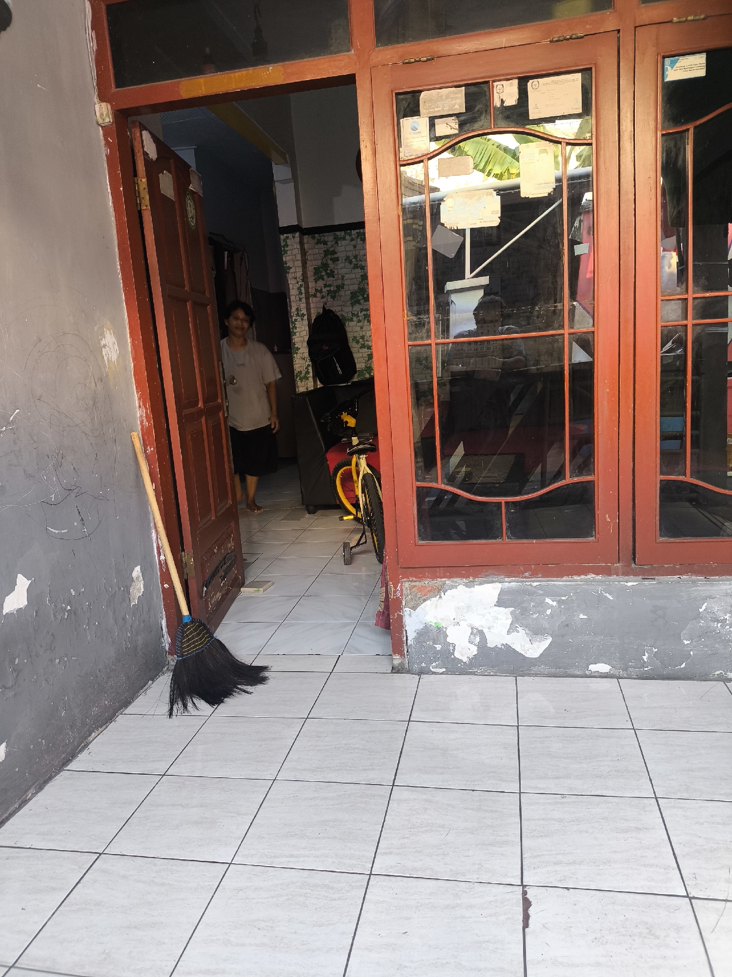 A medium shot captures a dimly lit, residential entrance in Citrodiwangsan, Indonesia. A woman, wearing a grey t-shirt and black skirt, stands in an open doorway. She appears to be facing away from the camera, looking into the interior of the house. The doorway is framed by a weathered red door and a larger red-framed window with multiple panes. The window is adorned with various documents and papers affixed to it.

To the left of the doorway, a broom with dark bristles and a wooden handle leans against a grey, textured wall. The wall shows signs of wear and peeling paint, revealing a rougher texture underneath.

The floor in front of the entrance is covered with large, light-colored square tiles, some of which are outlined by thin, dark grout lines. The tiles appear clean.

In the background, visible through the doorway and window, is an interior hallway with white tiled walls adorned with a green leafy pattern. A black backpack rests on a dark surface, possibly a couch or chair. A bicycle with a yellow frame and red wheel spokes is partially visible in the middle ground, positioned to the right of the doorway. The overall scene suggests a domestic, lived-in environment.